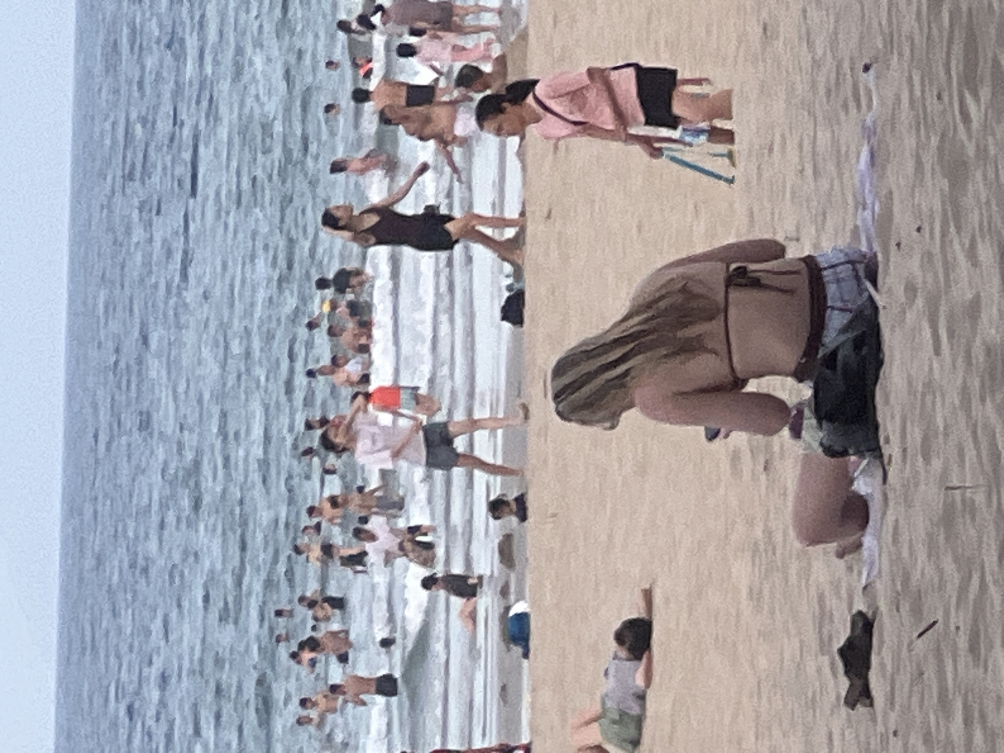 The image depicts a crowded beach scene in Da Nang, Vietnam, with many people swimming in the ocean and relaxing on the sand. The ocean water is blue and choppy, with waves rolling towards the shore. On the beach, there are several individuals in various states of leisure. In the foreground, a woman with long blonde hair is seated on the sand, facing away from the camera. To her left, a person in a green shirt and shorts lies on their back. Further back, a woman in a pink shirt and black shorts walks along the beach, carrying a blue bag. Numerous other people are visible in the water and on the sand, some standing, some sitting, and some playing. The overall atmosphere appears to be one of a busy, sunny day at the beach.