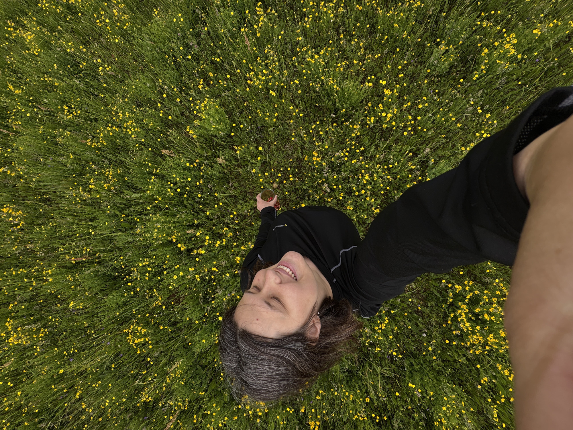 A high-angle, wide shot captures a person from the perspective of someone looking down. The person, positioned in the lower left quadrant of the frame, is seen from the chest up, smiling and looking upwards with eyes closed. They have shoulder-length brown hair with streaks of gray and are wearing a black long-sleeved shirt. Their right arm is extended towards the upper right, holding a small, clear glass with a red liquid and ice. The background is a dense field of bright green grass dotted with numerous small yellow wildflowers. The overall impression is a serene moment in nature. The location is Unknown, Unknown.