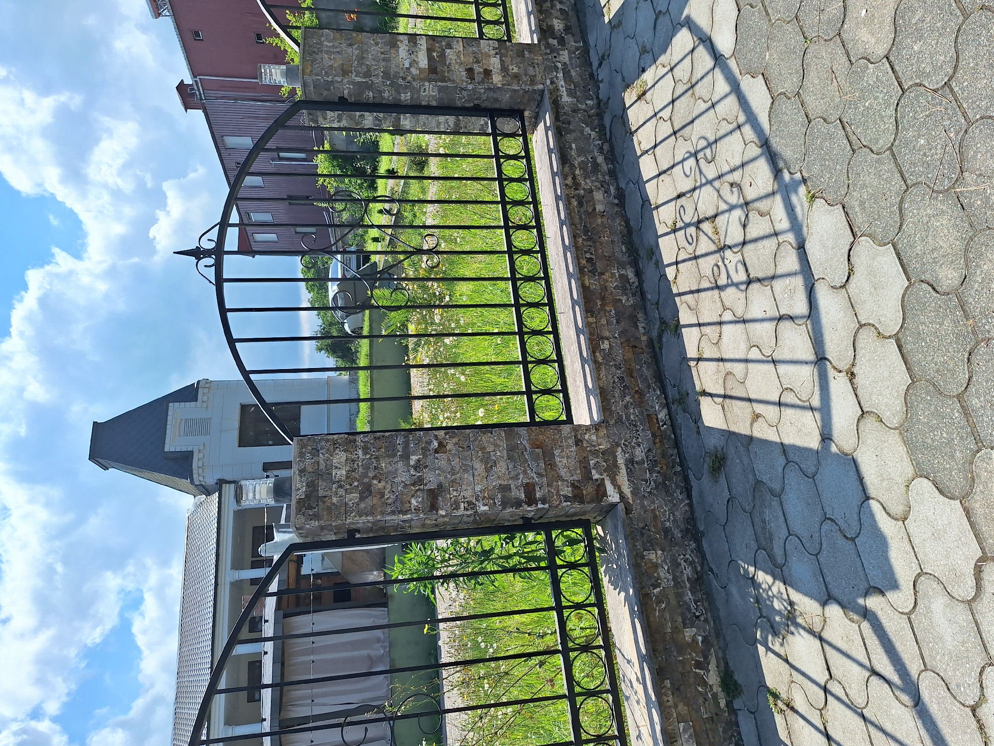 The image depicts a wrought-iron gate in front of a property, likely in Kobuleti, Georgia, under a bright, cloudy sky. The gate is supported by brick pillars and opens onto a grassy area with scattered white flowers. Beyond the grass, two buildings are visible: one is a modern, light-colored structure with a dark, steep roof, and the other is a larger, reddish-brown building with multiple windows.
The perspective of the photo is from ground level, looking upwards and slightly to the side. The sun is casting long shadows from the gate and railings onto the interlocking grey paving stones that form a walkway. The light suggests it is either morning or late afternoon. The weather appears to be fair, with a mix of sun and clouds. There is no discernible activity taking place; the scene is static and appears to be a view of the property entrance.
Notable details include the decorative metalwork on the gate, the texture of the brick pillars, and the pattern of the paving stones. The overall impression is one of a well-maintained residential or commercial property. No text is visible in the image.