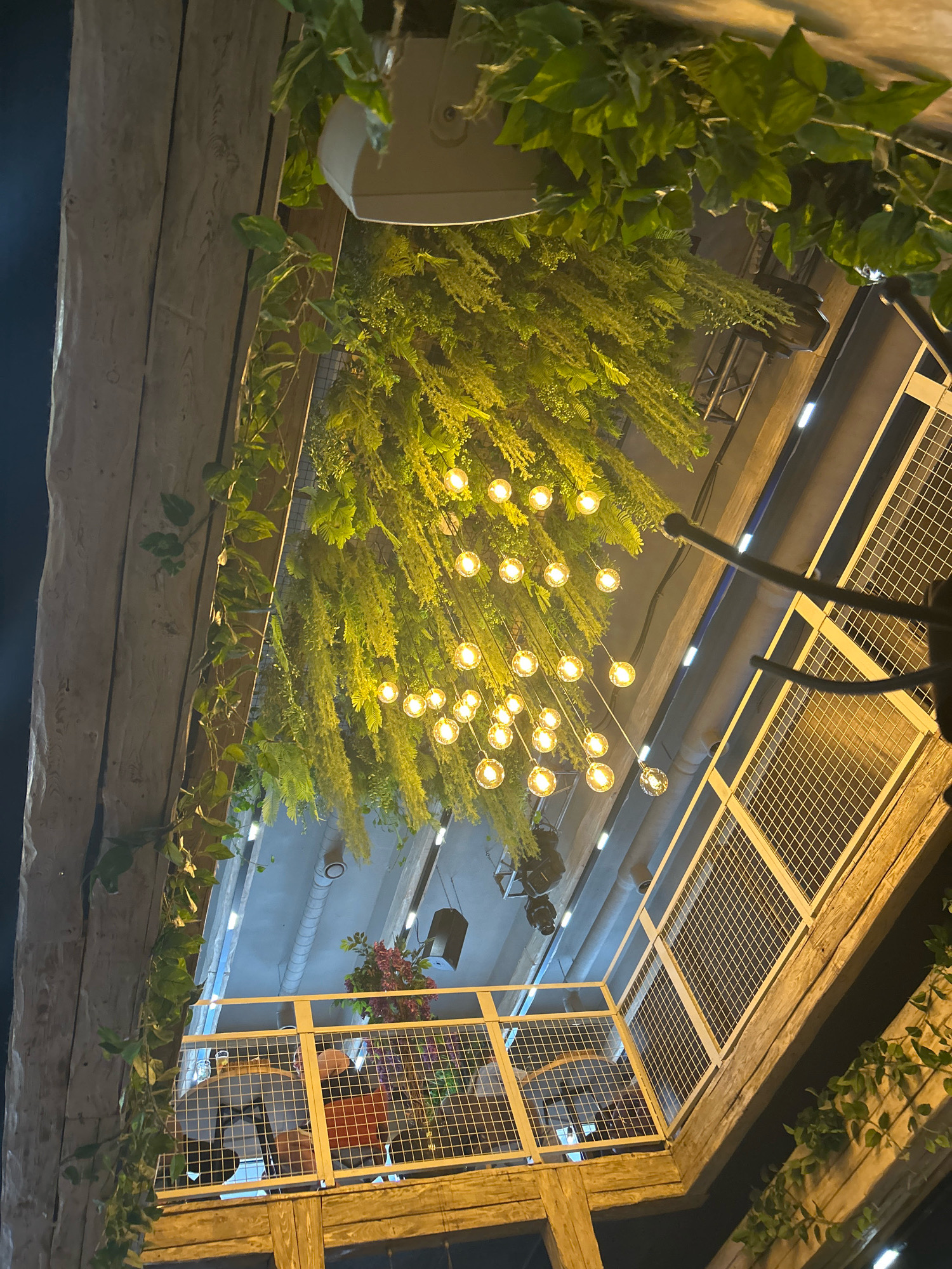 The image is an upward-angle shot of an outdoor seating area, possibly a cafe or restaurant, decorated with lush green hanging plants and a string of Edison-bulb lights. The lighting is warm and ambient, suggesting it is evening or nighttime. The seating area features metal railings with a mesh infill and wooden beams that form the structure. In the background, industrial elements like exposed pipes and lighting fixtures are visible. The overall aesthetic is modern and natural, with a focus on creating an inviting atmosphere. The scene appears to be in Moscow, Russia, as indicated by the available information.