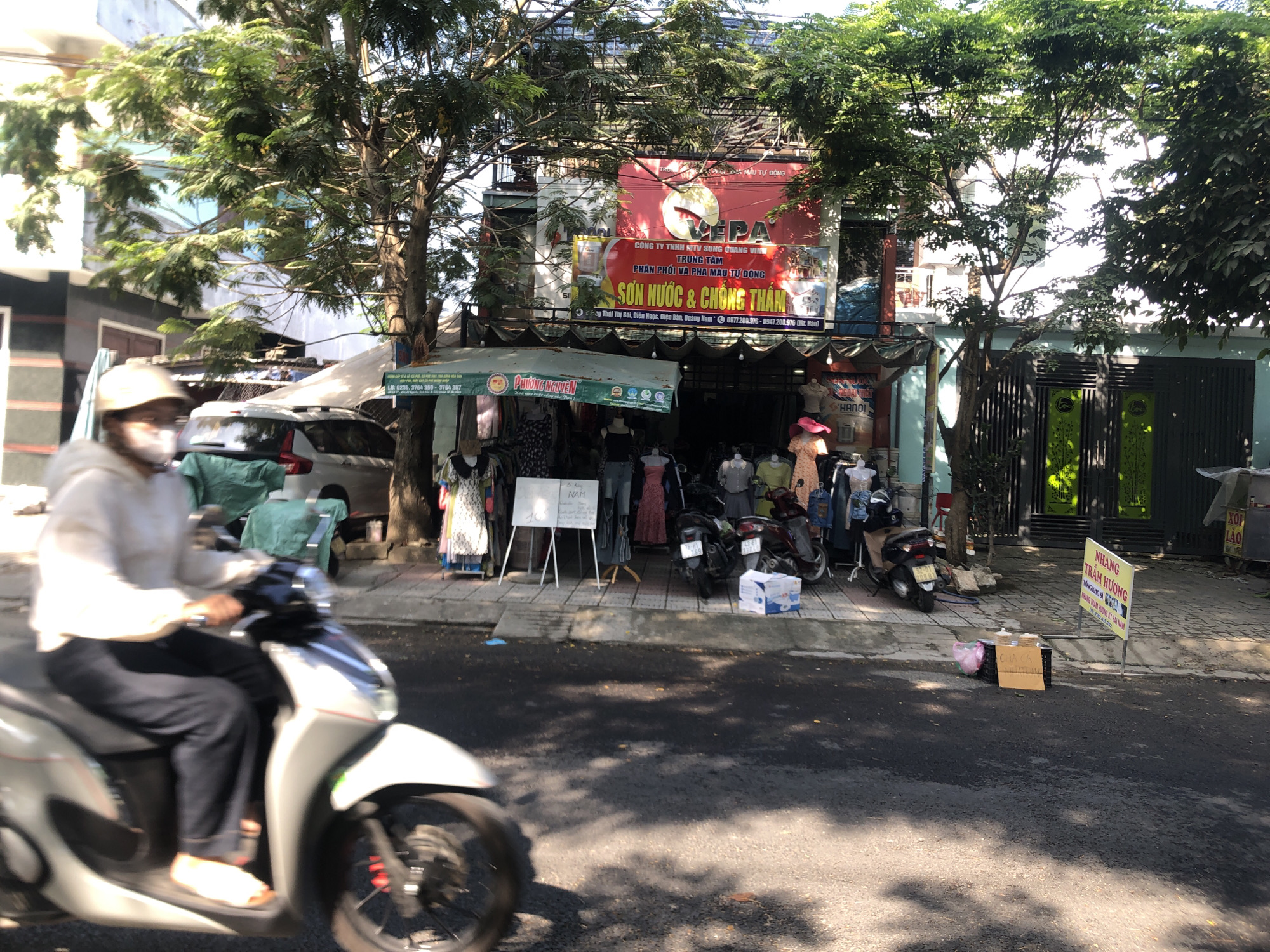 A street scene in Đà Nẵng, Vietnam, features a person riding a scooter on a sunny day. The rider, wearing a helmet and mask, is in motion on a light-colored scooter.
In the background, a two-story building displays a large red sign with Vietnamese text indicating a business named "VEPA" specializing in paints. Below the sign, an awning covers a storefront displaying clothing. Mannequins and clothing racks are visible on the sidewalk, along with a small whiteboard and parked motorcycles.
Sidewalk signage advertises products in Vietnamese, including incense. A parked car is partially visible behind the storefront. Trees line the street, providing partial shade. The street appears to be asphalt with scattered shadows.