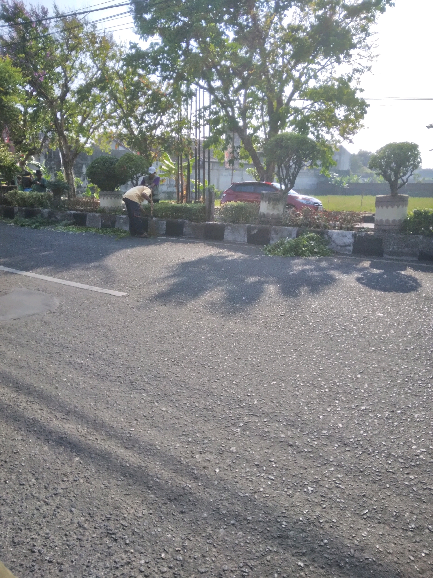 The image depicts a street scene in Purwokerto Kulon, Indonesia. In the foreground, a person in a light-colored shirt and dark pants is sweeping debris, likely leaves and plant trimmings, from the side of the road. A red car is visible behind a low decorative wall, further down the street. Several trees and ornamental plants are present, suggesting a well-maintained public area. The lighting indicates it is daytime, with shadows cast on the asphalt road. The overall environment appears to be a typical urban or suburban street in Indonesia.