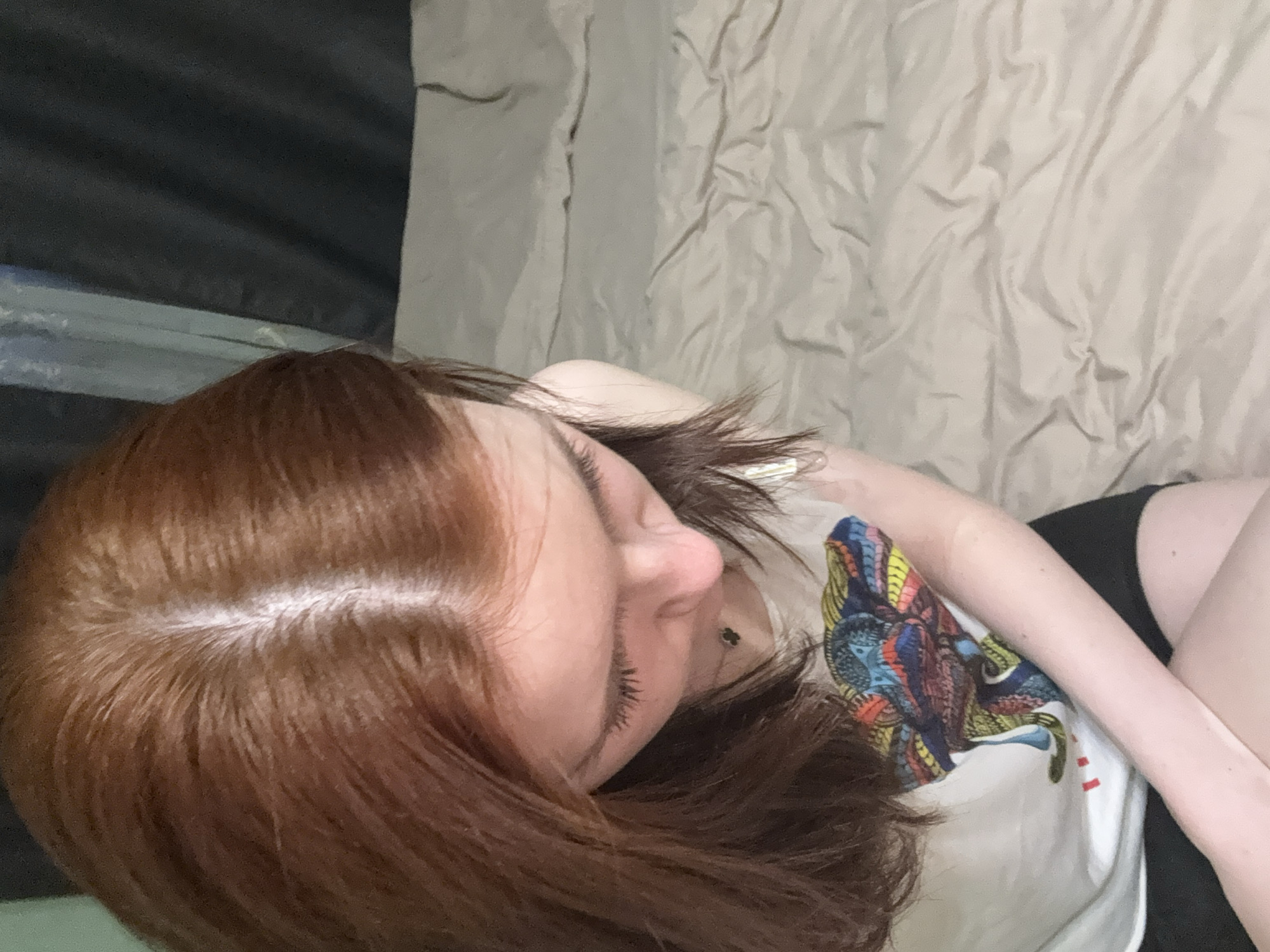 The image shows a person lying down on a bed. The person has reddish-brown hair with a visible scalp parting. They are wearing a white T-shirt with a colorful graphic design. The background consists of wrinkled beige bedding and a darker, possibly black, object or surface in the upper left corner. The location is Unknown, Unknown.