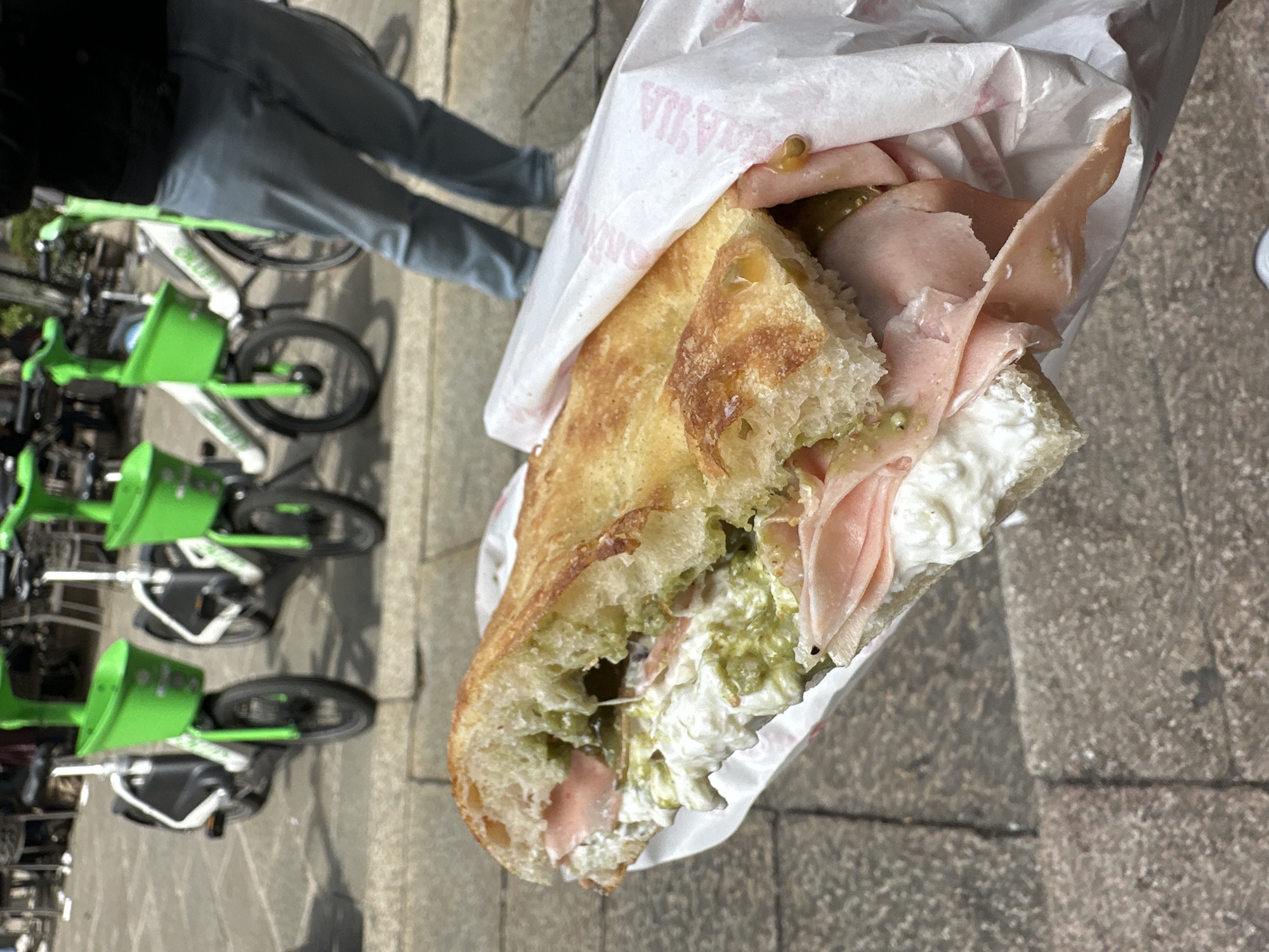 A close-up, slightly rotated photograph captures a person holding a large, generously filled sandwich wrapped in white paper, set against an urban background in Milan, Italy. The sandwich, appearing to be made with focaccia or a similar rustic bread, is split open to reveal its abundant fillings. These include several thin slices of light pink cured meat, a creamy white spread (possibly a soft cheese or mayonnaise), and a chunky green sauce or spread, with visible green olive pieces.
The white paper wrapping, creased around the sandwich, has faint red text, where parts of "VIA" and "ITALIA" are discernible, hinting at its origin.
In the background, a person wearing a dark jacket and grey trousers is visible from the waist down, standing on a grey paved surface. To their right, a row of bright green and white shared bicycles, identifiable as Lime bikes, are neatly parked. The setting suggests a bright, clear daytime outdoors, likely on a street or pedestrian area. The image focuses on the sandwich as if it's about to be eaten, capturing a moment of a casual meal in the city.