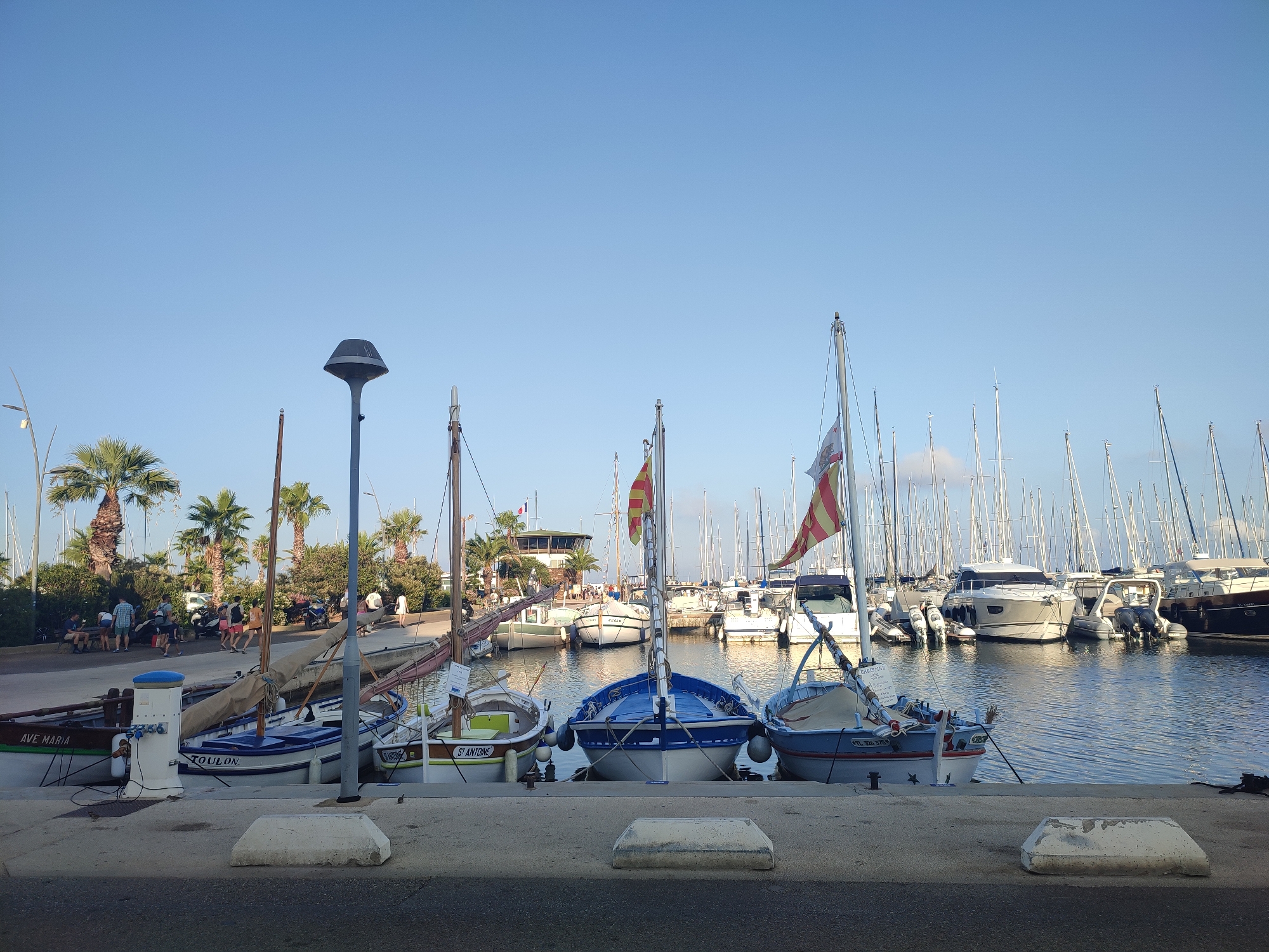 The image depicts a sunny day in a harbor, likely in Hyères, France, with a clear blue sky. The foreground shows a concrete walkway with several large, rough, white concrete blocks. Beyond the walkway, a row of boats is docked. The closest boats are smaller, traditional-looking vessels, one of which has "AVE MARIA" painted on its side. Another boat displays "TOULON" in blue lettering on its hull. A white electrical post is situated between these two boats.
Behind these smaller boats, a multitude of sailboat masts rise into the sky, indicating a larger marina. Some of these sailboats have flags flying, including one with red and yellow stripes. In the background, more substantial yachts are docked, and further back, a building with a distinctive observation deck or control tower is visible, surrounded by palm trees and other greenery. Several people are scattered along the waterfront, some walking, others sitting, and a few are near the boats. There are also parked motorcycles and a white vehicle visible in the middle ground. The water in the harbor is calm, reflecting the blue of the sky and the masts of the boats.