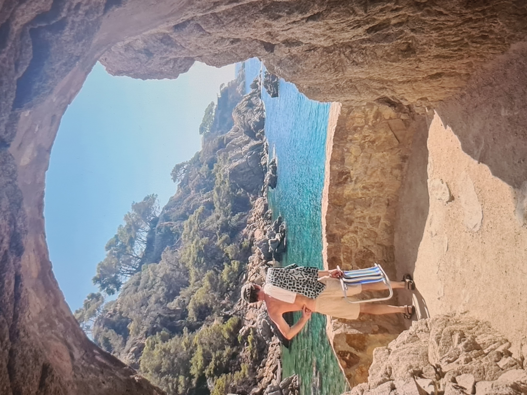 The image presents a picturesque coastal scene viewed from within a rocky cave or archway. A man stands in the foreground, his back to the camera, looking out at the breathtaking expanse of the sea and coastline. He is dressed in light-colored shorts and a short-sleeved top, carrying a folded blue and white striped beach chair in one hand and a patterned black and white bag or towel draped over his shoulder.
The man is positioned on a flat, sandy-toned rocky floor inside the cave, which frames the outdoor vista with its rough, textured brown and tan rock walls. Beyond the cave opening, a clear, vibrant turquoise sea stretches out under a bright blue sky. The water is calm, revealing hints of rocky formations beneath its surface. The coastline is rugged and steep, covered in dense green vegetation, primarily pine trees, adding to the natural beauty of the setting.
The scene suggests a warm, sunny day, likely during the daytime, with the bright light illuminating the man and the stunning natural environment. The man appears to be enjoying the view or perhaps looking for a spot to relax on the beach or shore. No visible text is present in the image.