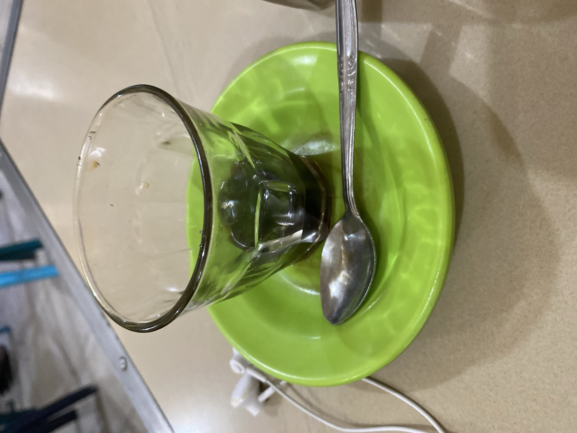 A close-up, eye-level shot shows a single-serving of dark liquid in a clear glass, sitting on a vibrant green saucer. A silver spoon rests on the saucer to the right of the glass, and a white earphone cord snakes from the left edge of the frame, disappearing under the saucer. The scene is set on a neutral-toned tabletop, with blurred blue and gray furniture visible in the background. The lighting suggests an indoor setting, possibly a cafe or restaurant in Lhokseumawe, Indonesia.