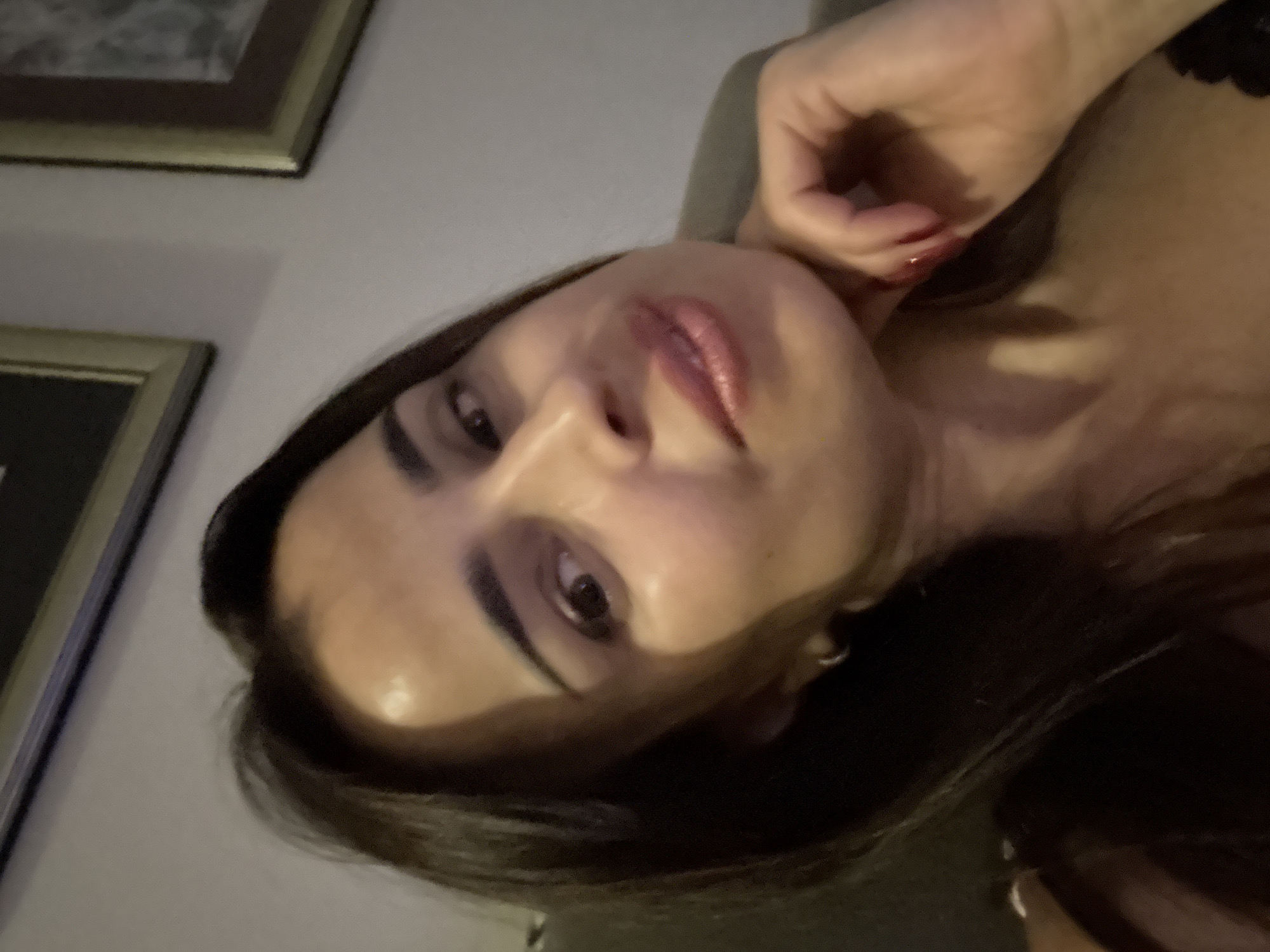 A medium shot, close-up selfie of a woman with long, dark hair and light skin is captured from a slightly low angle. She is resting her chin on her right hand, which has red nail polish. Her eyes are looking slightly to the side, and her mouth is slightly open. The background is out of focus, but appears to be an indoor setting with framed pictures on a wall. The lighting is dim and appears to be artificial.