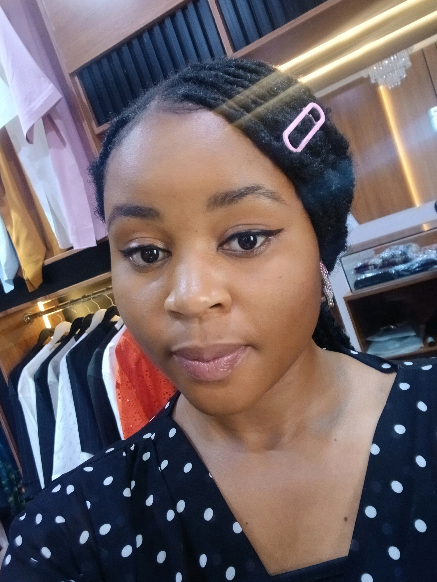 A medium shot, taken from a slightly low angle, features a young woman looking directly at the camera. She has dark skin, dark braided hair styled with a pink clip, and is wearing a black and white polka dot top. The background reveals a closet with neatly hung clothes on hangers, including a pink shirt on the left, and shelves stocked with items on the right. The lighting in the scene appears to be artificial, possibly from overhead lights that create a glow.