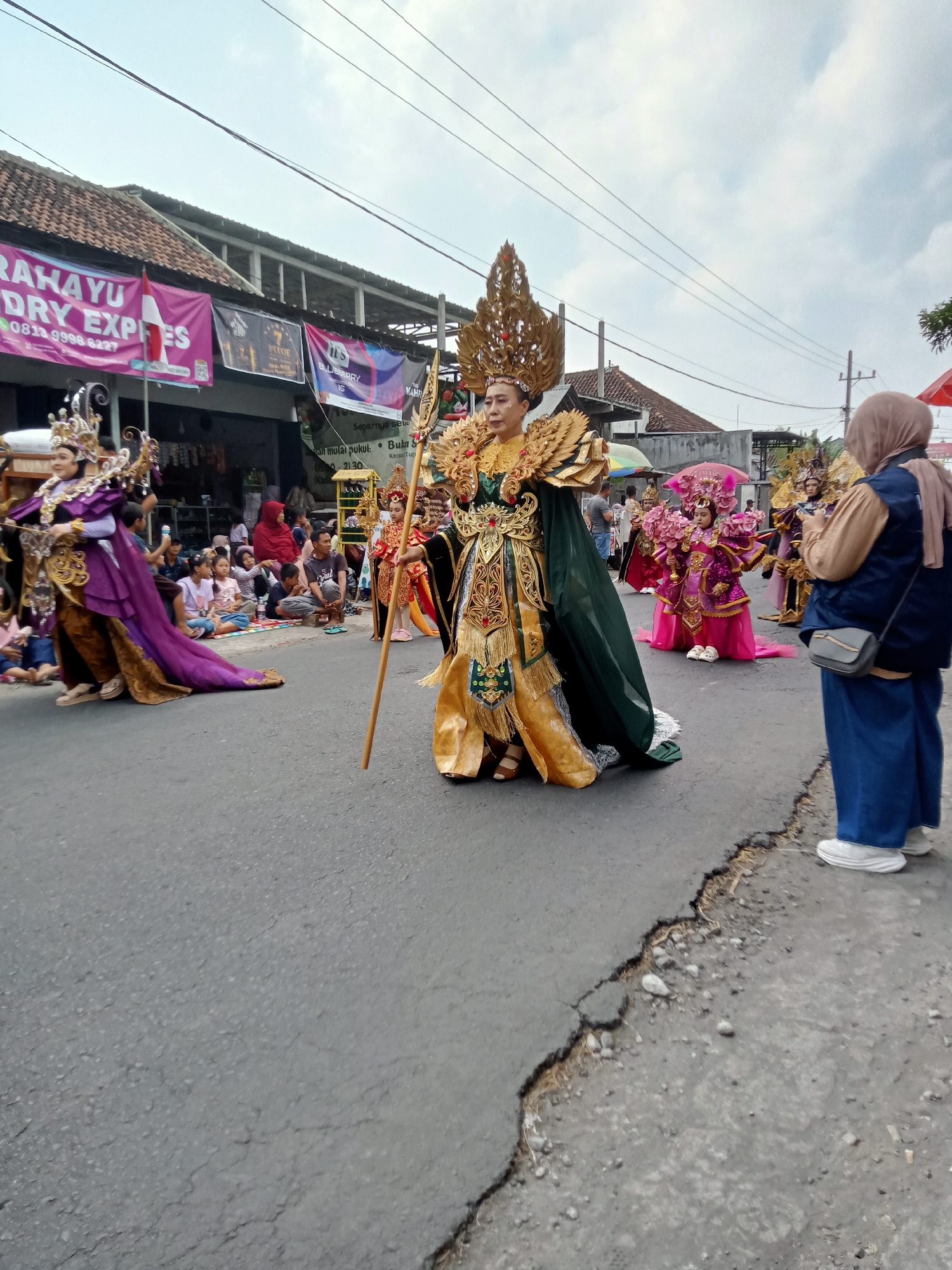 This is a medium shot, eye-level, outdoor photograph taken on a street in Simo, Indonesia, featuring a parade. The foreground is dominated by the cracked asphalt road. The middle ground shows several people in elaborate costumes participating in what appears to be a cultural procession.
On the left, a person in a regal purple and gold outfit with a long train is walking, facing towards the right. To their right, the central figure of the parade is a person adorned in a magnificent green and gold costume, complete with a towering, intricate golden headdress and a golden scepter. This individual is the primary focus and is facing forward, slightly to the right.
Behind these central figures, other participants are visible, including a child in a bright pink and gold costume and a person in a dark outfit with gold accents. The background reveals a row of shops with banners, including one that reads "RAHAYU DRY EXPRES". Spectators are seated on the side of the road, watching the parade. A woman in a hijab and blue denim pants is standing on the right side of the road, holding a phone and looking towards the parade. The sky is overcast.