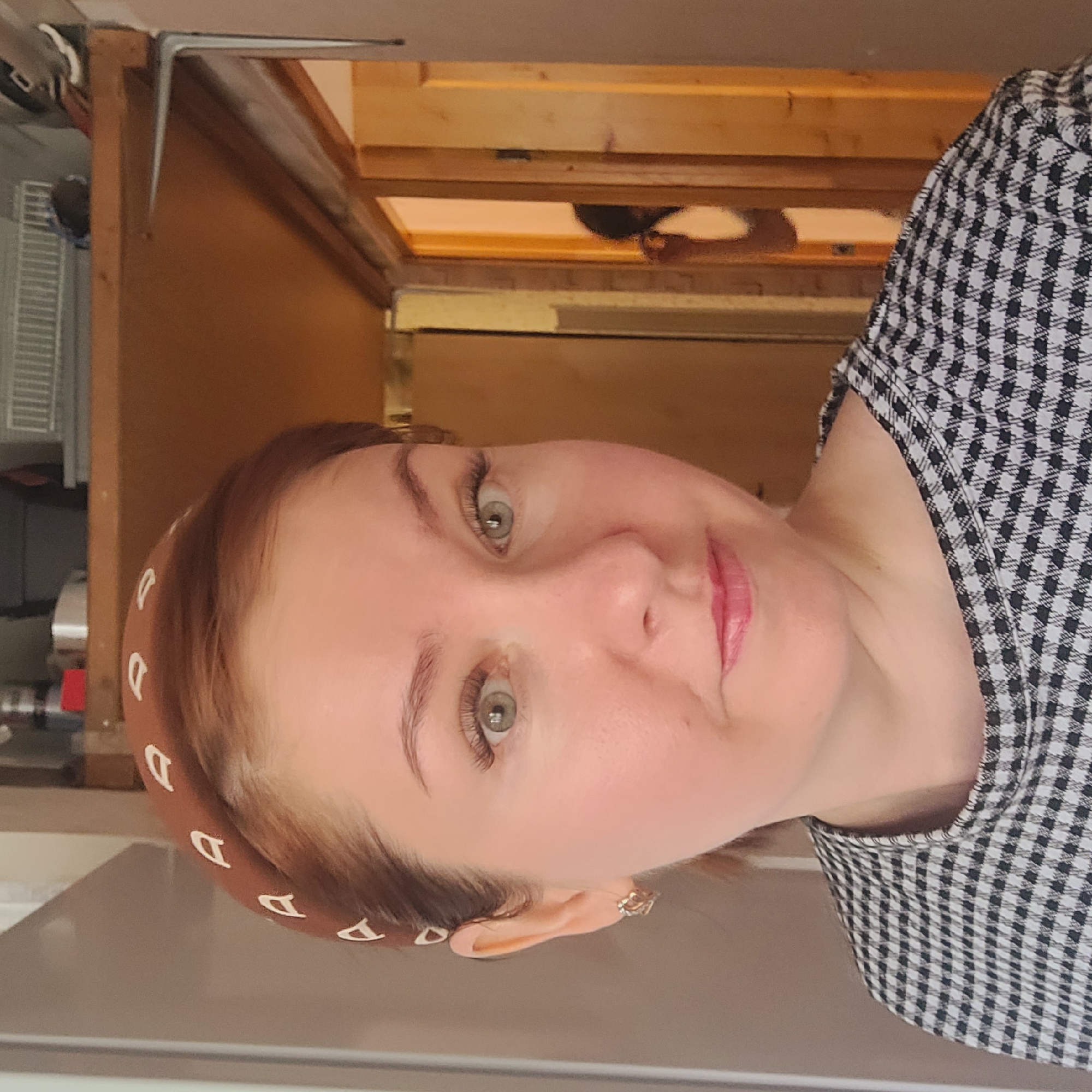 A close-up portrait shows a fair-skinned woman with green eyes and light brown hair wearing a headband with white "D" patterns and a black-and-white checkered shirt. She is indoors, with a wooden cabinet visible behind her. The location appears to be a residential area, specifically the city of Unknown, Unknown, judging by the domestic interior.