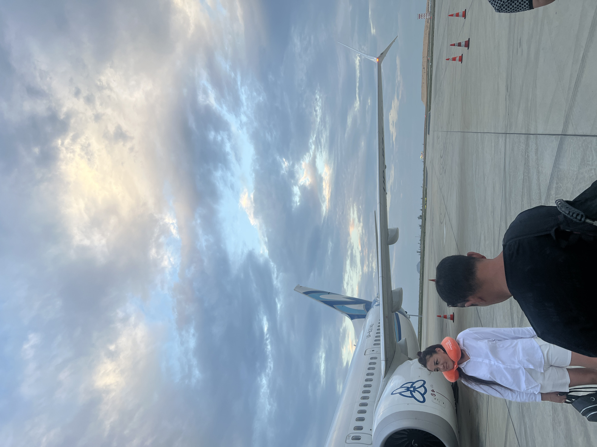 This media file depicts an outdoor scene on an airport tarmac, likely in Bac Cam Ranh Ward, Vietnam. The time of day appears to be either sunrise or sunset, indicated by the soft, warm light filtering through a sky filled with dramatic, layered clouds.
The primary subjects are two individuals standing next to a large passenger airplane. On the right, an adult figure, seen from behind, wears a dark shirt and checkered shorts. To their left, a person, likely female, is dressed in a white shirt and white shorts, sporting an orange neck pillow. She is positioned close to the aircraft's engine, looking downwards.
The dominant object is a white passenger airplane with blue and gray livery accents. Its tail fin displays a distinct blue and white design. The aircraft registration "VP-B3740" is clearly visible on the fuselage and tail. A stylized blue logo, resembling interconnected rings or a 'B', is present on the engine cowling. The massive wing of the aircraft extends into the distance, with a light visible on its wingtip.
The ground is a grey concrete tarmac with expansion joints. In the mid-ground, three orange and white traffic cones are spaced out on the tarmac. The activity suggests passengers waiting to board or having just disembarked from the plane, underscoring a travel event. The overall mood appears calm and neutral.