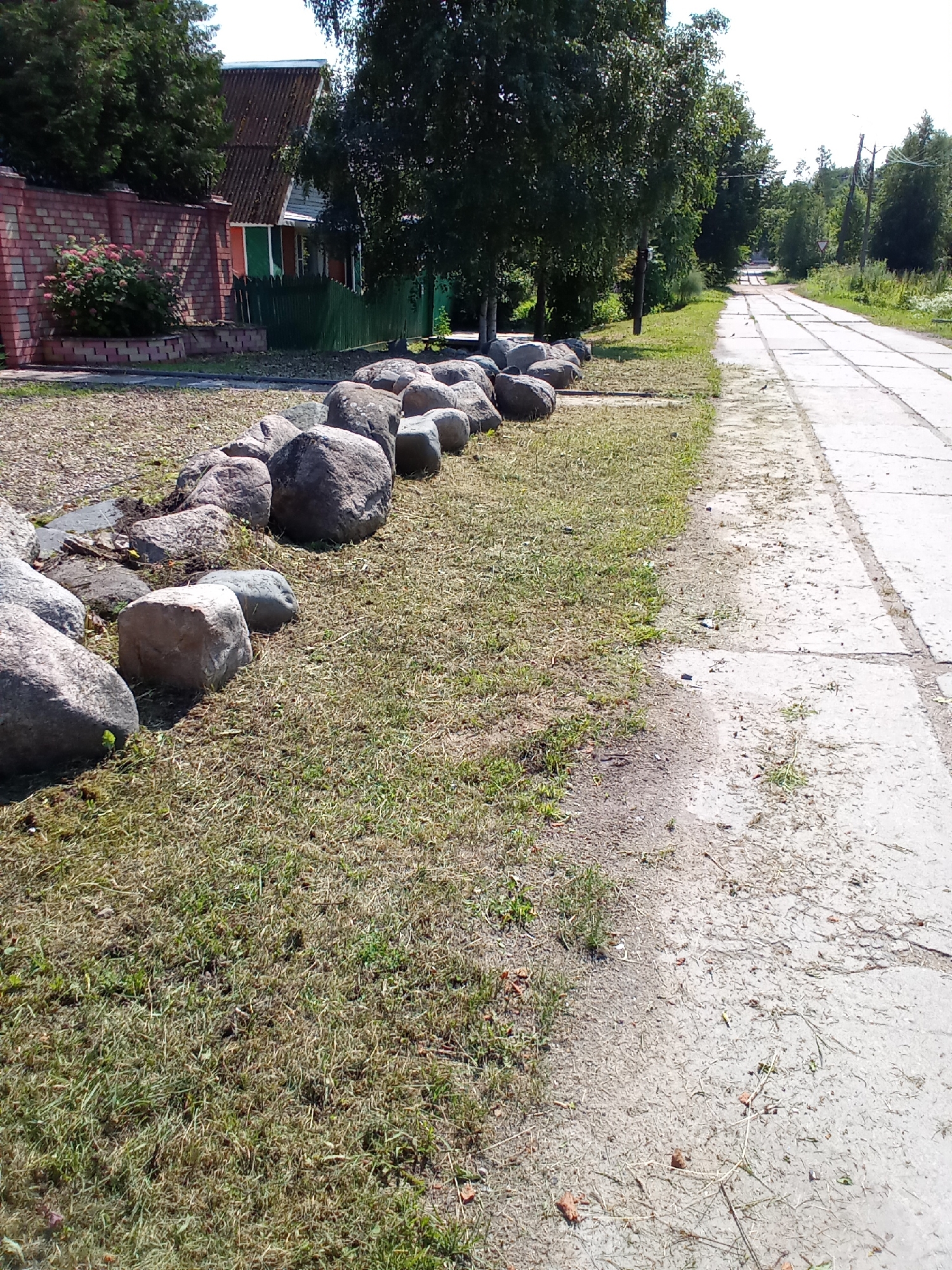 The image shows a paved pathway or road with concrete slabs, bordered by a grassy area. A line of large, rough-hewn rocks is arranged along the edge of the grass, separating it from what appears to be a gravel-covered area or the side of the road. Beyond the rocks and grass, there is a brick wall, a fence, a house, and trees. The pathway extends into the distance, curving slightly and lined with more trees on either side. The scene appears to be in a rural or suburban setting in Belozersk, Russia, likely a residential street or a path in a park. The lighting suggests it is daytime, with clear skies. No people are visible in the image.