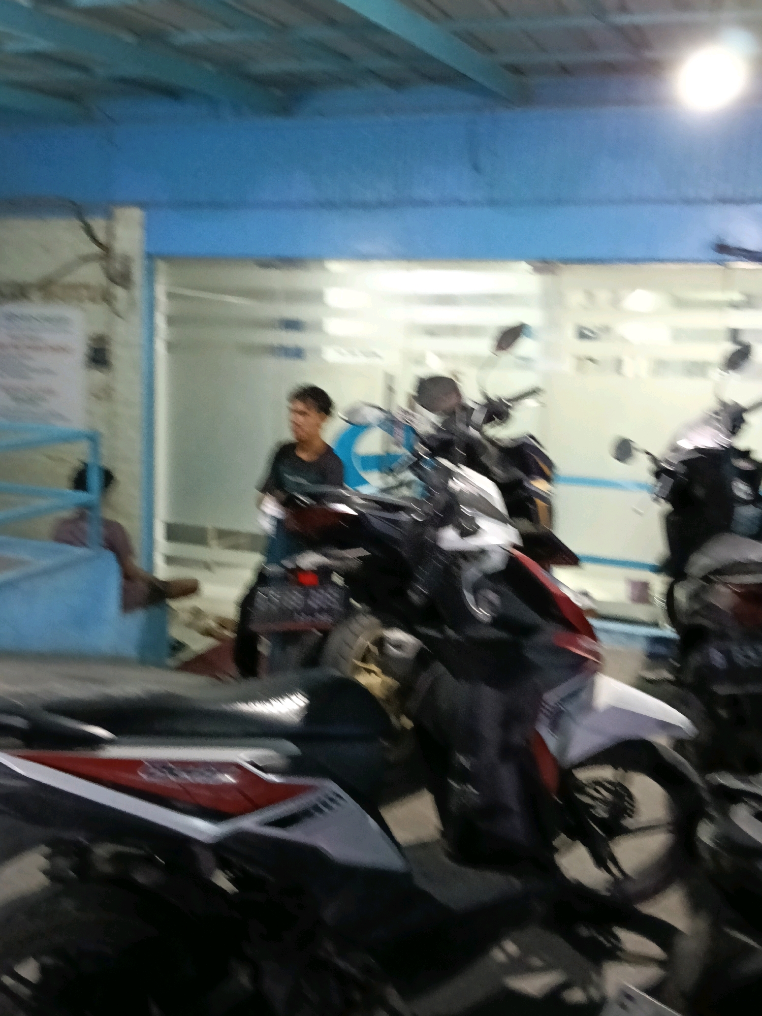 In Dadap, Indonesia, a man wearing a dark t-shirt and dark pants stands near a motorcycle. The scene is blurred, with the background showing a building with blue trim and glass windows. Several other motorcycles are parked around the man and the one he is standing next to. The lighting suggests it is daytime.