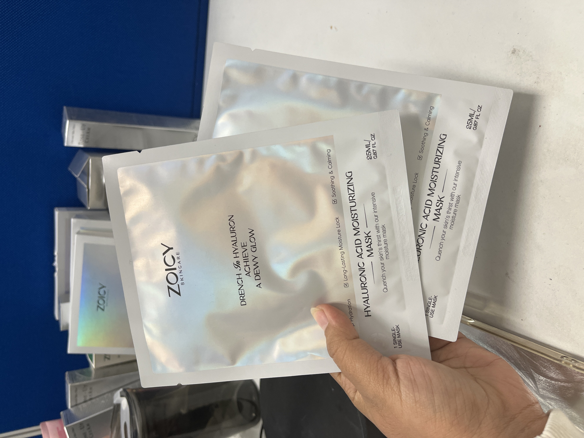 A close-up shot shows a person's hand holding two ZOICY Hyaluronic Acid Moisturizing Masks. The masks are packaged in shimmering, iridescent foil sachets. The top mask has text that reads "DRENCH IN HYALURON ACHIEVE A DEWY GLOW" and features checkboxes for "Hydration," "Long-Lasting Moisture Lock," and "Soothing & Calming." Below this text, the mask is further identified as "HYALURONIC ACID MOISTURIZING MASK" with the description "Quench your skin's thirst with our intensive moisture mask" and a volume indication of "25ML / 0.87 FL. OZ." The second mask, partially obscured, is identical in packaging and text. The background is blurred, showing a stack of other cosmetic product boxes, including one with "ZOICY" printed in holographic lettering. The environment appears to be an indoor setting, possibly an office or retail space, with a blue textured wall visible in the upper right portion of the image. The scene is well-lit, emphasizing the reflective quality of the mask packaging.