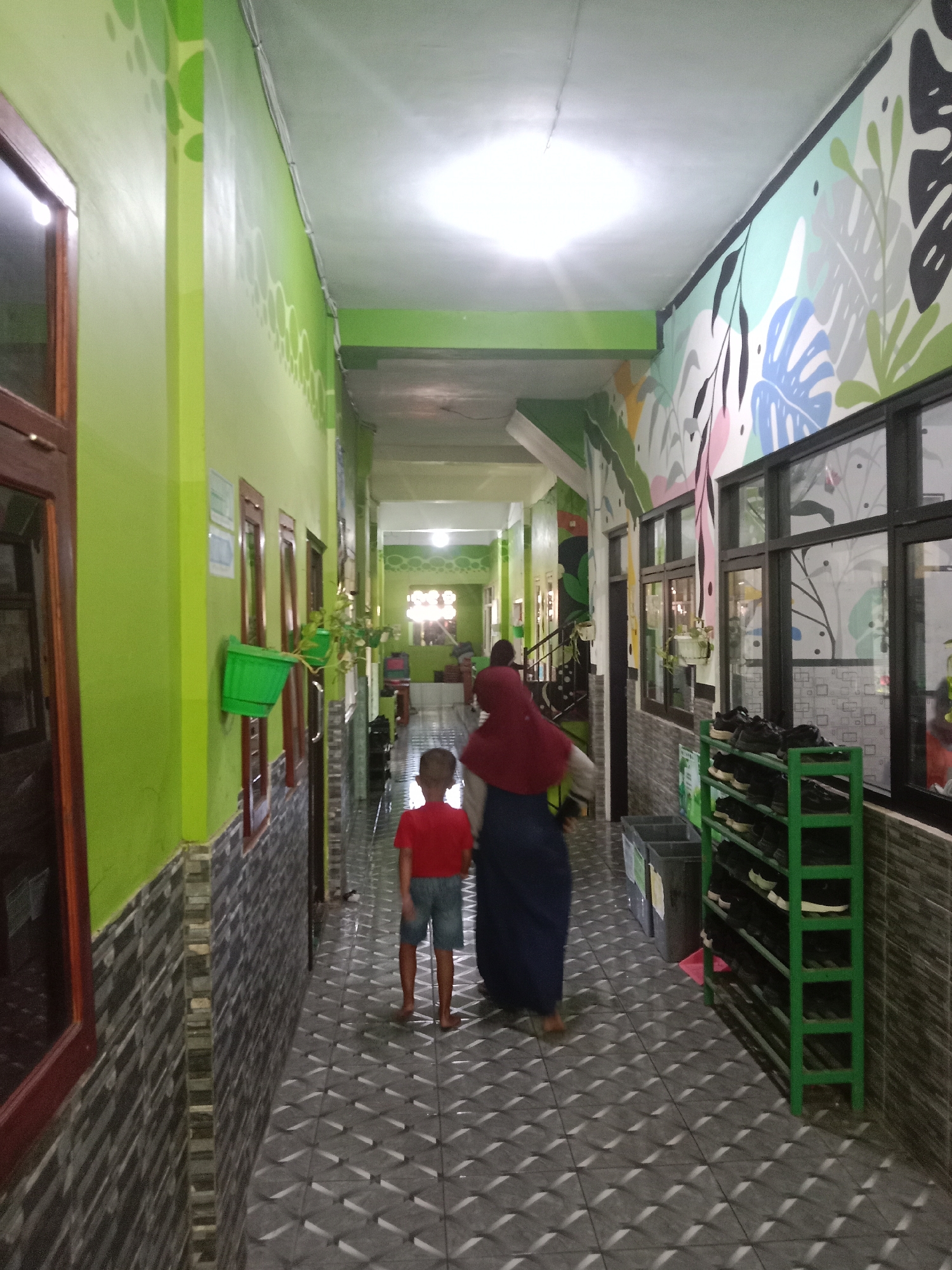 The image depicts a long, brightly lit hallway with green walls adorned with stylized plant murals on one side and windows on the other. The floor is tiled with a grey geometric pattern that reflects the light. On the left side of the hallway, there are several wooden-framed windows and a green planter box attached to the wall. On the right side, there is a green shoe rack filled with black shoes, and two grey trash bins.
Two individuals are walking down the hallway, away from the camera. The person in front, on the right, is a child wearing a red t-shirt and blue shorts. The person behind the child is an adult wearing a maroon headscarf and a dark blue skirt. The hallway leads towards a brightly lit area at the far end, where more people can be seen. The overall impression is that of an indoor public space, possibly a school or community center.
The provided context indicates the location is in the city of Probolinggo, Indonesia. This suggests the hallway is part of a building within that Indonesian city.
