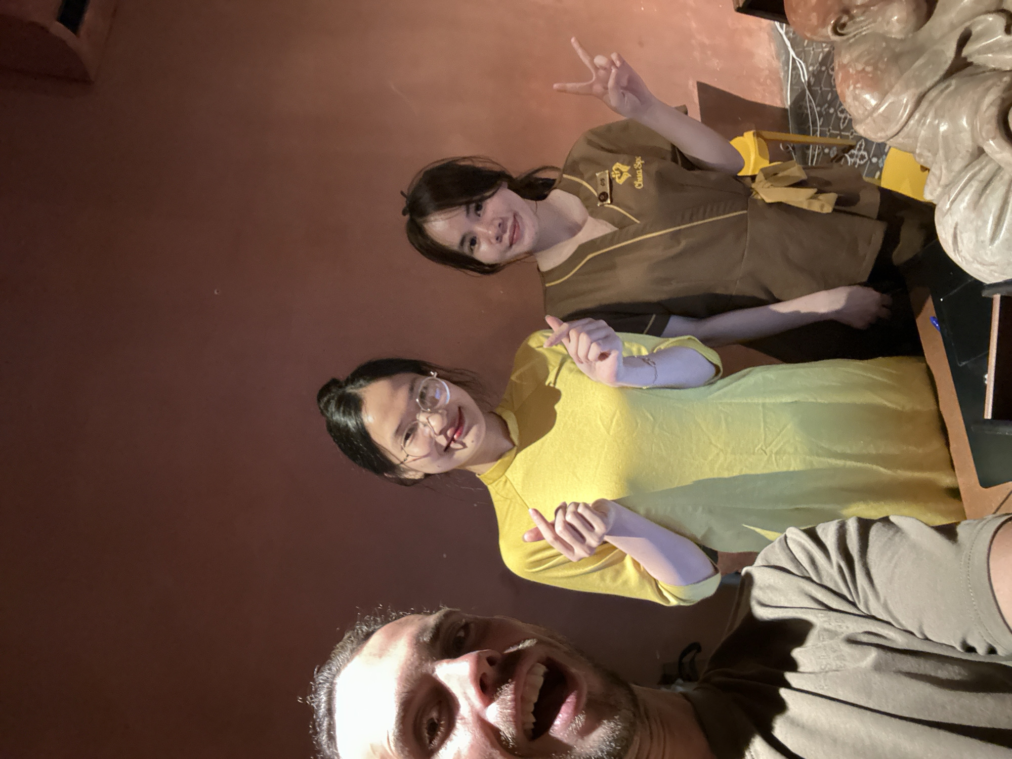 The image depicts a group of three individuals posing for a selfie in what appears to be an indoor establishment with dimly lit, warm-toned walls. In the foreground on the left, a Caucasian man with short, dark, curly hair and a beard is smiling widely, his mouth open. He is wearing a plain, dark greenish-brown t-shirt. To his right, a young Asian woman stands in the center, wearing a bright yellow, long-sleeved garment. She is wearing glasses and has dark hair pulled back, with her hands held up in front of her chest, forming a heart shape with her thumbs and index fingers. Next to her, on the right, another young Asian woman is also posing. She is wearing a brown uniform with yellow trim, possibly a spa or salon uniform, which has a logo on the left chest that reads "Chua Spa." Her dark hair is pulled back into a ponytail. She is making a peace sign with her right hand. The background wall is a solid, muted reddish-brown color. To the right of the women, a bright yellow chair is visible. In the bottom right corner, the top of a large, intricately carved light-colored stone statue, possibly of a seated figure with a serene expression, is visible. The lighting suggests an indoor setting, possibly a spa or massage parlor in Đà Nẵng, Vietnam, given the presence of the "Chua Spa" logo, a common spa name in Vietnam. The overall atmosphere is casual and friendly, with the individuals engaged in a playful interaction.