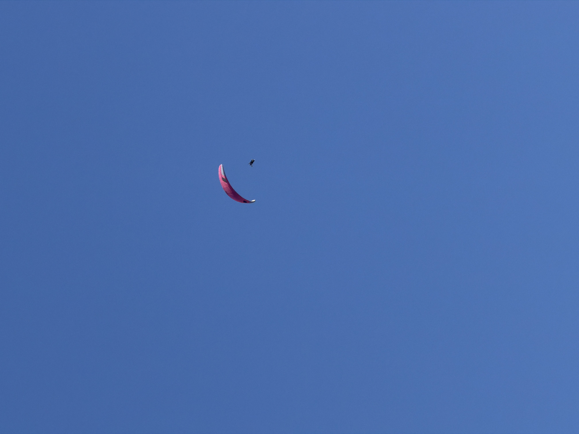 A paraglider is flying in the sky over Boreti, Montenegro. The paraglider is red and the sky is clear blue.