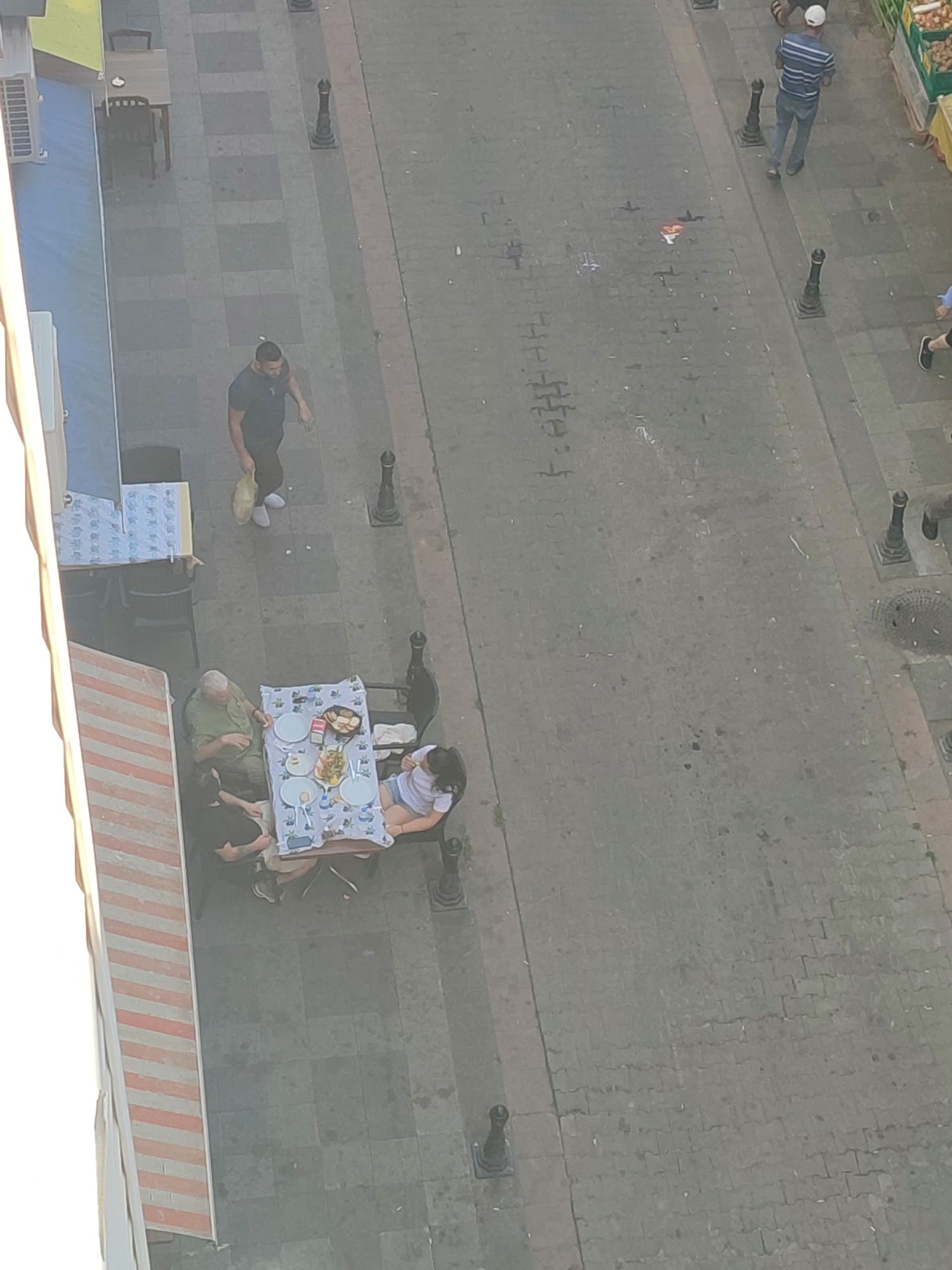 The image, taken from an elevated perspective, depicts a street scene in Kadıköy, Turkey. In the foreground, a group of three people are seated at a table outdoors. The table is covered with a patterned tablecloth and set with plates and food, suggesting a meal is in progress. One individual, wearing a green shirt, is seated facing left. Another individual, wearing black, is seated to their left. A third individual, wearing a white top and blue shorts, is seated to the right. Adjacent to this table, another table is visible, covered with a blue and white patterned cloth. Further up the street, a man in a dark t-shirt and dark pants is walking and carrying a yellow bag. In the upper portion of the image, another person in a striped t-shirt and jeans is walking away from the camera. The street is paved with interlocking stones. Various bollards are positioned along the sidewalk. The overall setting appears to be an outdoor seating area of a cafe or restaurant on a street.