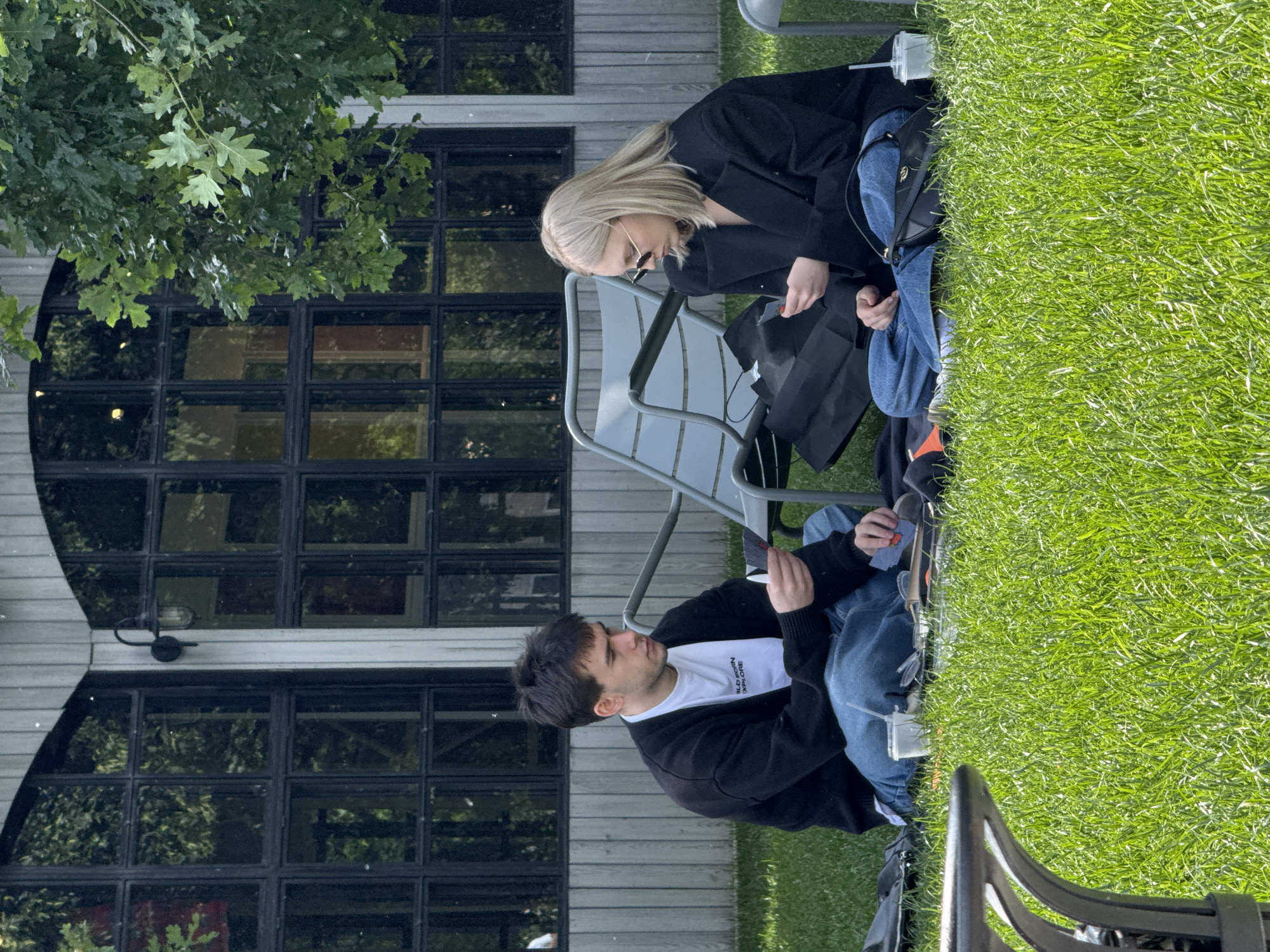 A young man and woman are sitting on the grass in front of a building with large windows. The man, on the left, is wearing a black cardigan over a white t-shirt and blue jeans. He is holding playing cards in his hands. The woman, on the right, is wearing a black jacket and blue jeans. She is also looking at playing cards in her hands. There are black bags and a plastic cup on the grass between them. A grey lounge chair is behind them. The building in the background appears to be a modern structure with wooden siding and dark-framed windows. Trees are visible to the left of the building. The scene is set in a park-like area with well-maintained green grass. The overall impression is of a casual outdoor gathering, possibly in a park or a courtyard in Saint Petersburg, Russia.