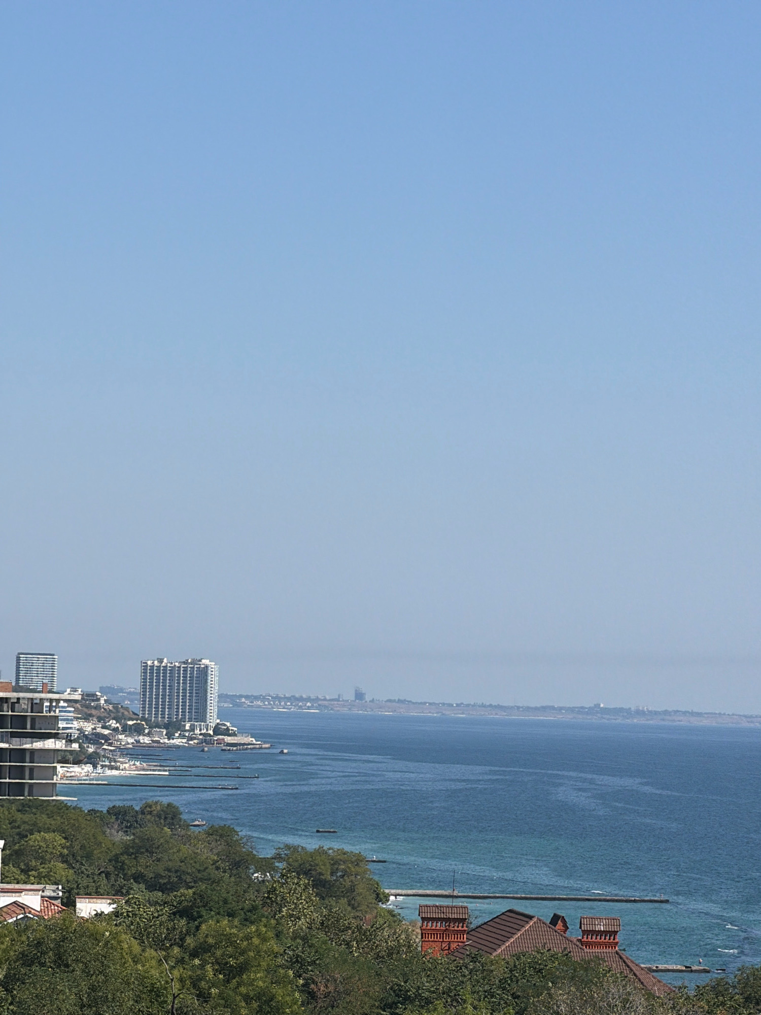 The image depicts a wide view of the Odesa coastline on a clear day. In the foreground, lush green trees cover a hillside, with the tiled roofs of several buildings visible amongst them. As the view moves towards the middle ground, a row of coastal buildings and high-rise apartment complexes line the shore. One prominent white high-rise building stands out, alongside other multi-story structures and what appear to be partially constructed buildings. The sea stretches out in the background, its waters a deep blue, with several long, narrow piers extending out into the water. A distant coastline is visible on the horizon, with faint structures suggesting further development. The sky above is a clear, pale blue, indicating a sunny day. There are no people visible in the frame, but several small boats are scattered across the water.