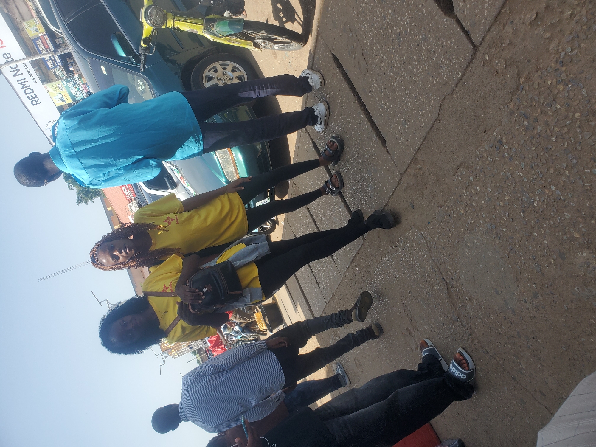 The image depicts a street scene in Jos, Nigeria, under a bright, clear sky. In the foreground, four individuals are visible, with two women in yellow t-shirts standing close together, one holding a black backpack. To their left, a man in a blue long-sleeved shirt and dark pants stands facing away from the camera. Further left, a man in a striped shirt is also partially visible. Behind the main group, a green car is parked, and a yellow motorcycle is positioned to the right of the car. The ground is paved with concrete slabs, showing some cracks. In the background, buildings and other vehicles are present, suggesting an urban environment. The overall impression is a candid snapshot of people interacting on a city street.