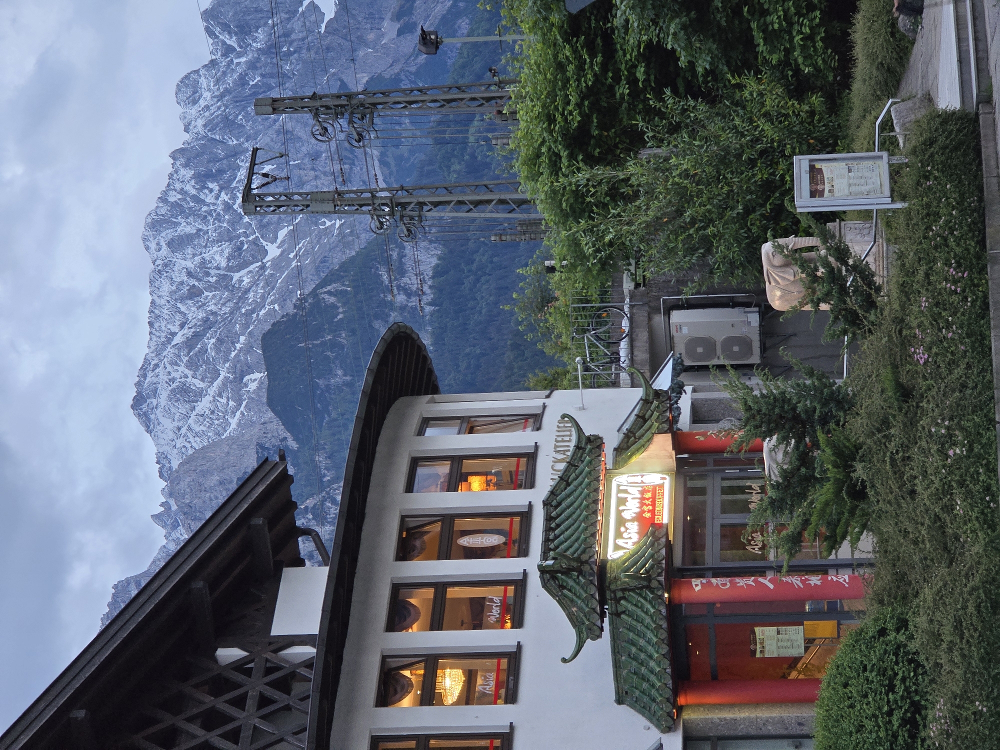 The image shows a building with an Asian-inspired architectural design, likely a restaurant, in Garmisch-Partenkirchen, Germany. The building features a white facade, with a prominent green, tiled roof over the entrance, adorned with red columns. A neon sign above the entrance reads "Asia World EDELGRÜSSE BUFFET". The building is situated against a backdrop of a lush green forest and imposing, snow-capped mountains. In the foreground, there are bushes and a pathway. To the right of the building, there are tall electrical pylons with overhead wires, suggesting proximity to a railway line. A bicycle is leaning against a railing on a raised walkway. A stone elephant statue stands near the entrance, and a poster is displayed on a stand. The sky is overcast, indicating potentially cool weather.