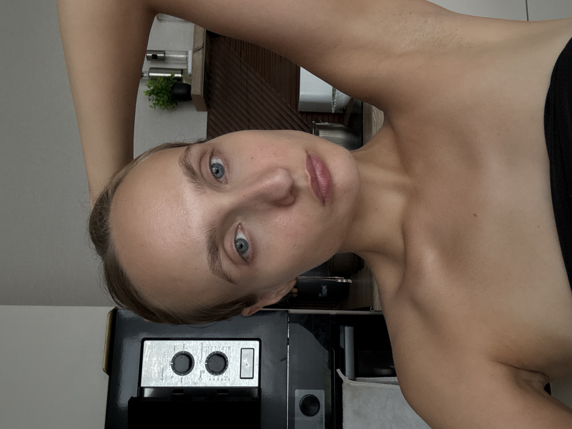 A young woman with light skin, light brown hair, and blue eyes is pictured in what appears to be a kitchen. She is looking directly at the camera with her right arm raised behind her head. Her left hand is not visible. She is not wearing any clothing above the shoulders. In the background to the left is a black microwave oven. To the right, there is a shelf with two small plants, a salt and pepper shaker, and a white container. The woman is wearing a black garment that covers her torso below the chest. The lighting suggests it is daytime.