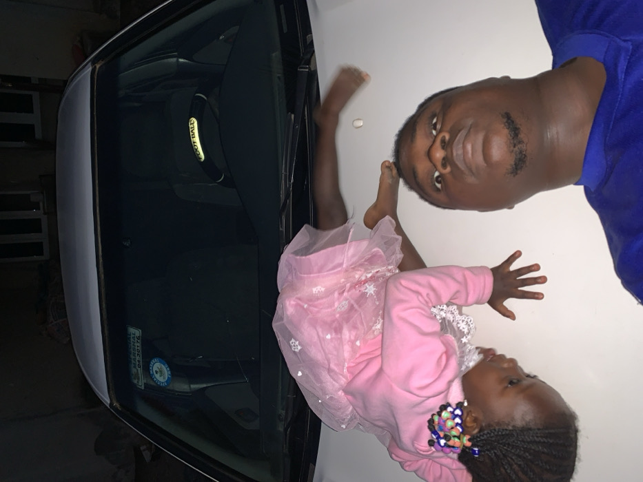 An upside-down image shows a man in a blue shirt next to a white car. On the car hood lies a young girl wearing a pink dress with white snowflakes and a pink sweater. Her hair is braided and decorated with colorful hair ties. A black football-branded wristband is visible through the car's windshield. A sticker on the driver's side indicates a registration renewal date of 04-30176.