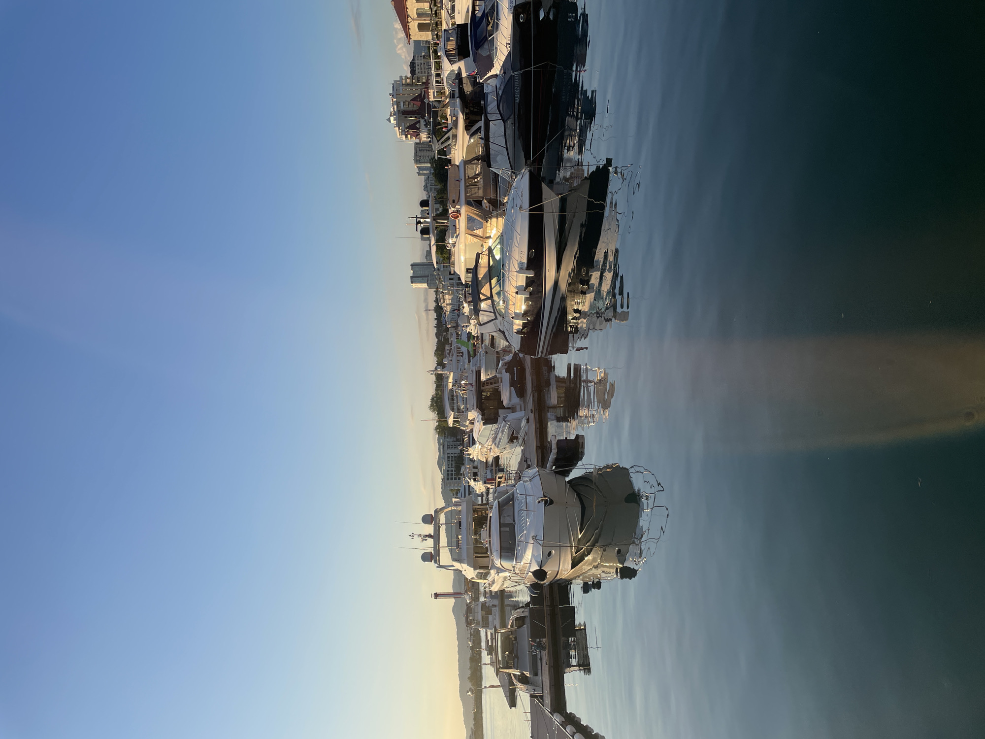 This is a photograph of a marina in Sochi, Russia, likely taken during the golden hour as the sun sets. The image depicts a collection of luxurious yachts and smaller boats docked along wooden piers. The water is calm and reflects the boats and the clear blue sky. In the background, there are buildings, including some with red roofs, and a lighthouse. The overall scene is serene and picturesque, conveying a sense of leisure and wealth associated with yachting.