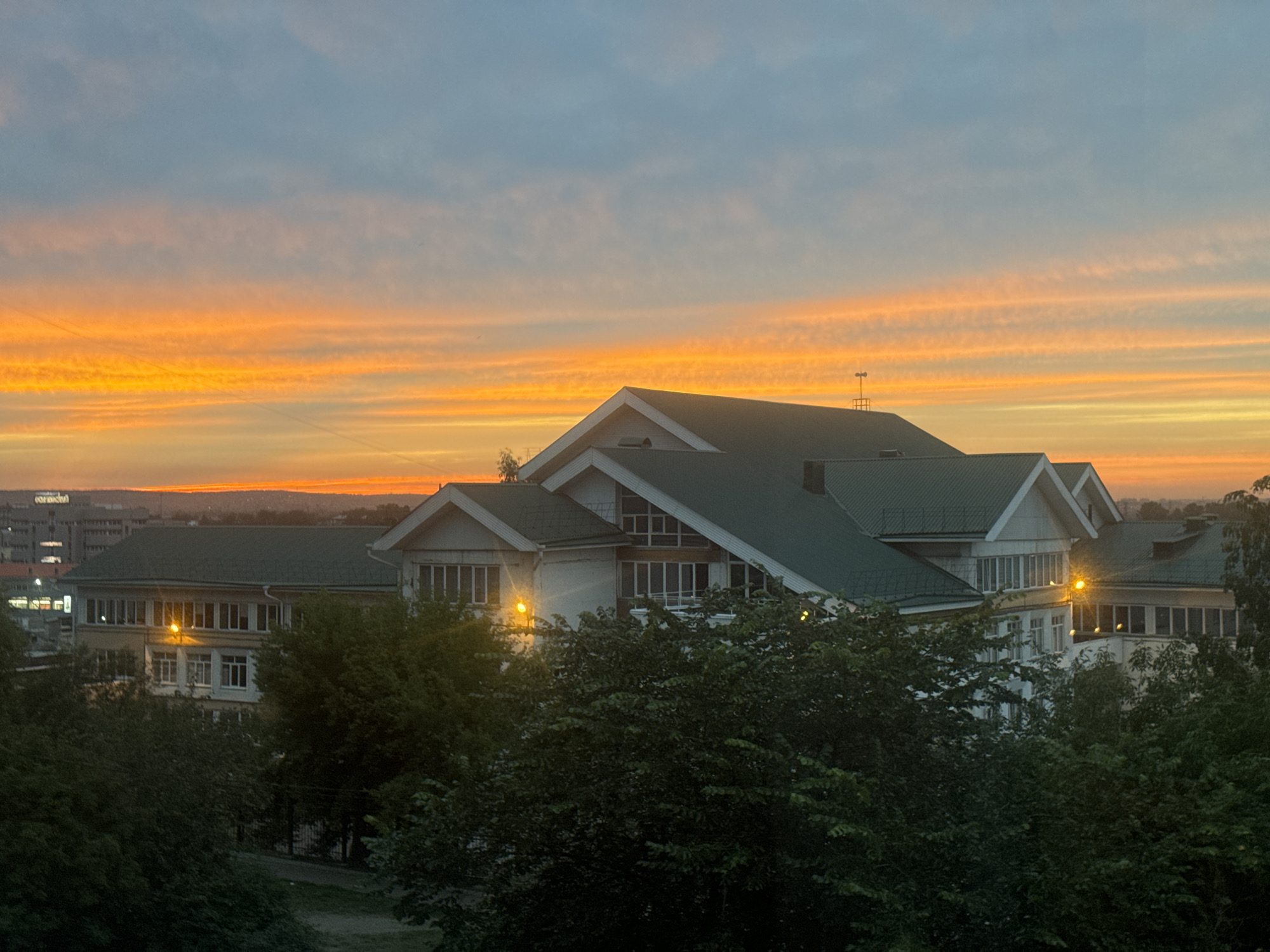 The image captures a serene sunset over a city, likely Irkutsk, Russia, given the context. The sky is ablaze with vibrant streaks of orange and yellow, transitioning to a soft blue towards the top, indicating the twilight hour. In the foreground, lush green trees partially obscure a multi-story building with a distinctive dark green roof. The building exhibits a modern architectural style with numerous windows. Streetlights are visible, casting a warm glow on the facade of the building and illuminating some of the windows. In the background, more urban structures are visible, suggesting a populated area. The overall scene evokes a sense of calm and natural beauty as day transitions into night.