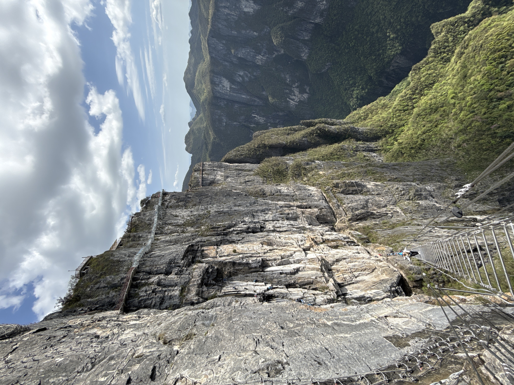 The image depicts a cliff face in Yongding District, China. A metal staircase is affixed to the rock face, and several people are climbing the stairs. Ropes are strung along the rock face, with metal rungs attached. The sky is visible above, with patches of blue and white clouds. A green, mountainous landscape can be seen in the background.