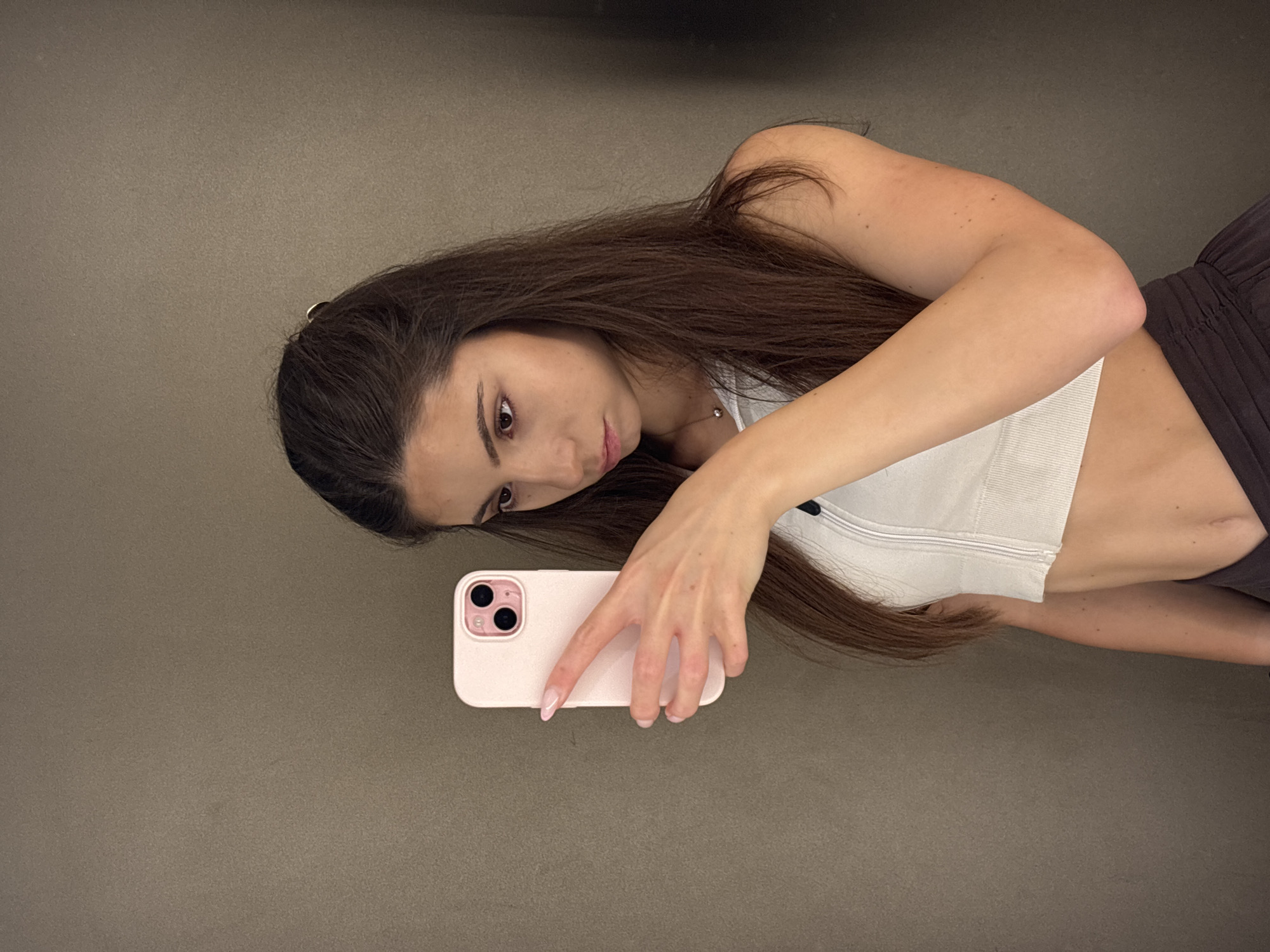 The image shows a young woman taking a selfie with her phone. She has long, dark brown hair and is wearing a white crop top and dark bottoms. Her phone is a light pink iPhone. The background appears to be a neutral-colored wall or floor. The lighting suggests it is daytime, but there are no external windows visible to determine the specific time of day or weather. The woman's expression is neutral. The setting is likely indoors, possibly a home or studio. There is no visible text in the image. The image orientation is portrait, and the woman is positioned in the center.