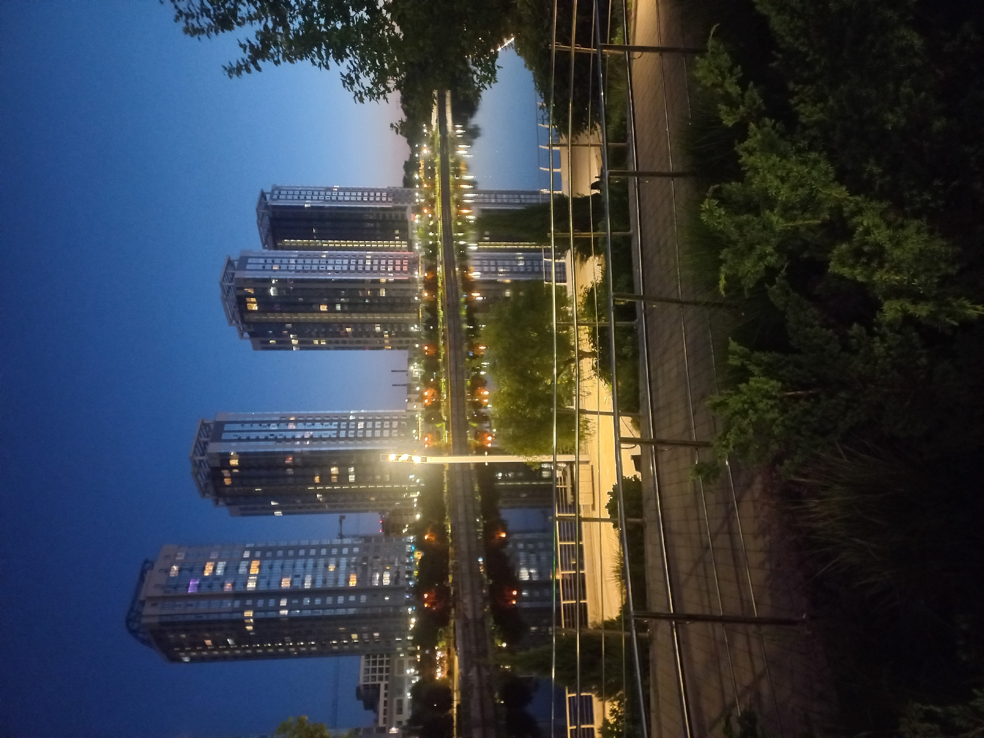 The image shows a night view in Kyiv, Ukraine, featuring modern architecture reflected in a body of water. The scene includes four tall residential buildings, with many windows lit, suggesting occupancy. The dark blue twilight sky contrasts with the buildings' lights. A waterfront pathway with a metal railing runs along the right side of the image, bordered by greenery, including trees and shrubs. The reflections of the buildings and lights are visible in the water. A light post stands along the pathway.