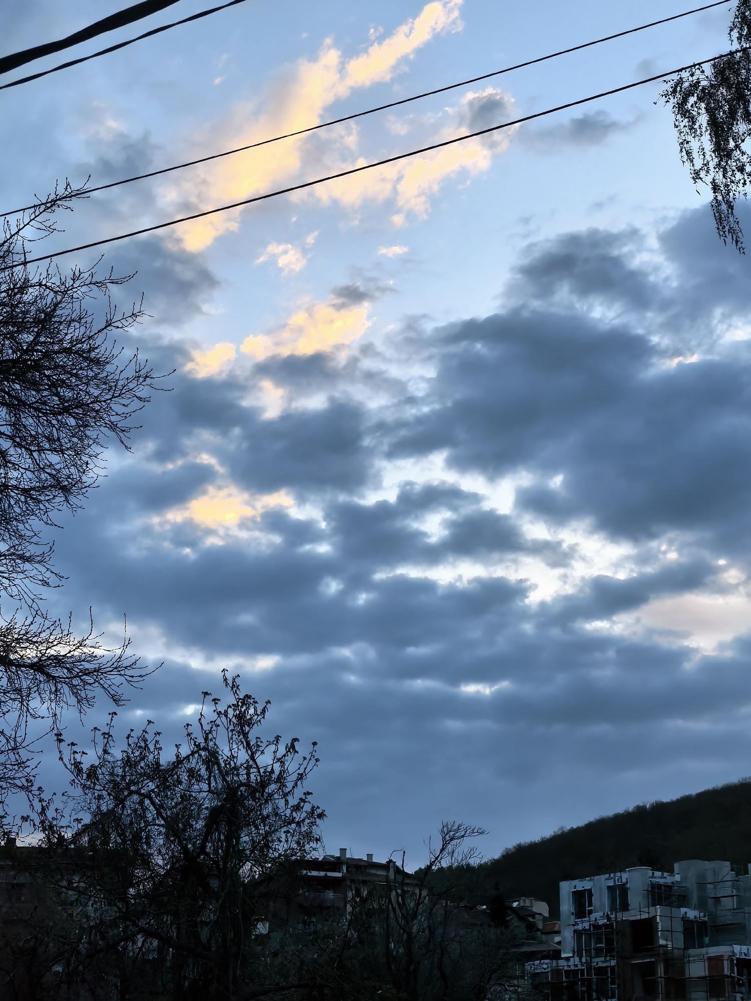 The image presents an outdoor scene under a cloudy sky in an urban environment. Power lines stretch across the upper portion of the frame. The clouds exhibit varying shades of grey and some are tinged with yellow, suggesting the presence of sunlight. Bare tree branches frame the sides of the image. The lower section of the scene reveals building structures, including one that appears to be under construction, indicating ongoing development in the area. A dark hill rises in the background, with trees silhouetted against the sky. The overall impression is one of urban life existing against a backdrop of natural elements. The location is the city of Unknown, Unknown.