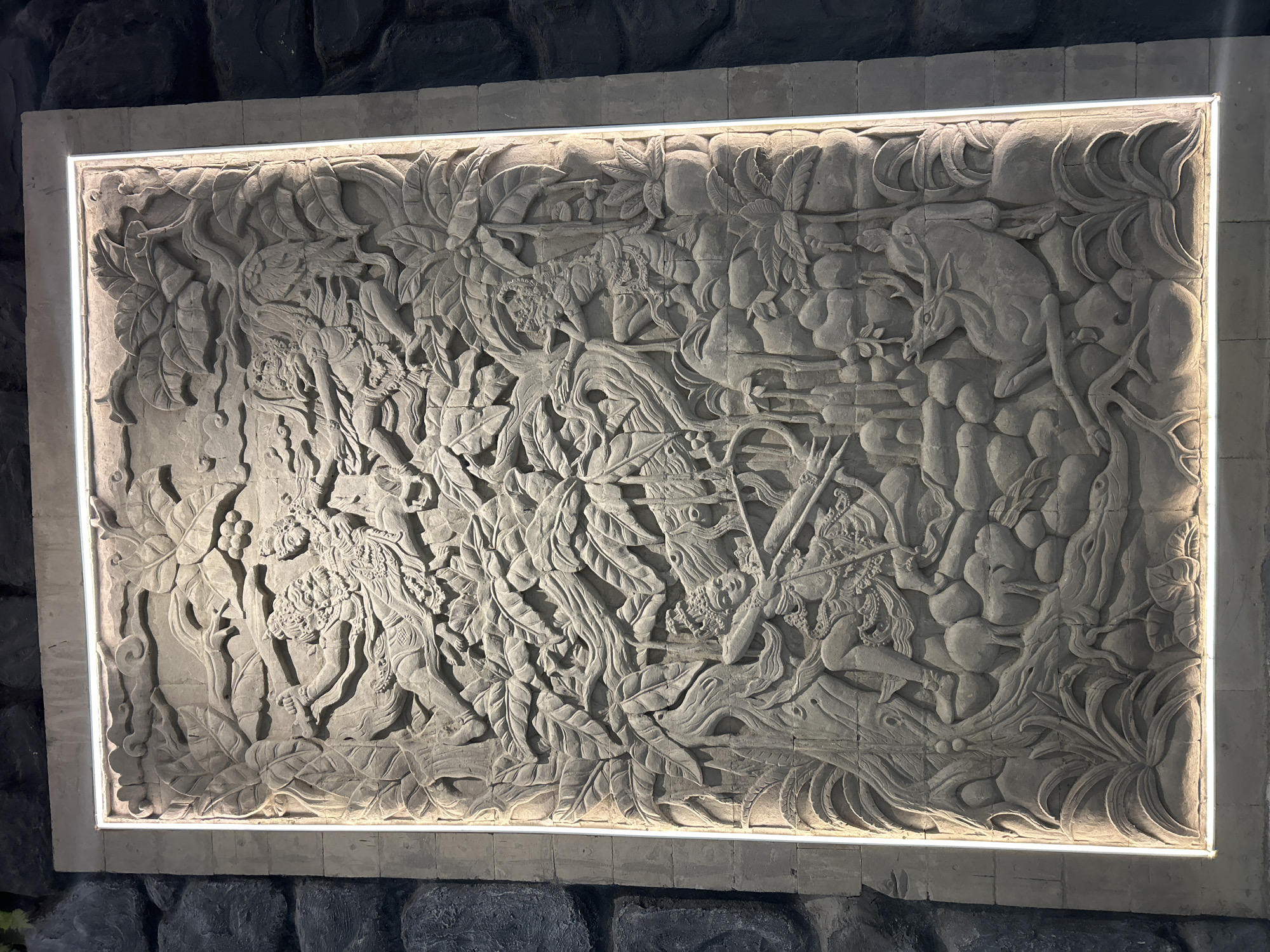 This is a detailed stone relief carving set against a textured dark stone wall, illuminated by white LED lights around its rectangular frame. The carving depicts a dynamic scene with multiple figures and elements, likely from a mythological or historical narrative.
In the upper left, a figure with a fierce expression and elaborate headdress is lunging forward, brandishing a weapon. Below and to the left, another figure, possibly a warrior, is aiming a bow and arrow. This figure is adorned with ornate jewelry and a crown. To the right of this archer, a distressed deer-like animal is depicted, possibly being hunted or in peril. Other figures, some with intricate headdresses and clothing, are also present, engaged in various actions, suggesting a conflict or a chase.
The environment is rich with carved foliage, including large leaves reminiscent of tropical plants, and intertwined branches. The overall composition is dense and layered, creating a sense of depth and movement within the stone. The carving is executed in a traditional Balinese style, characterized by its detailed ornamentation and expressive figures.
This carving is located at the entrance of the Bali Museum (Museum Bali) in Denpasar, Bali, Indonesia.