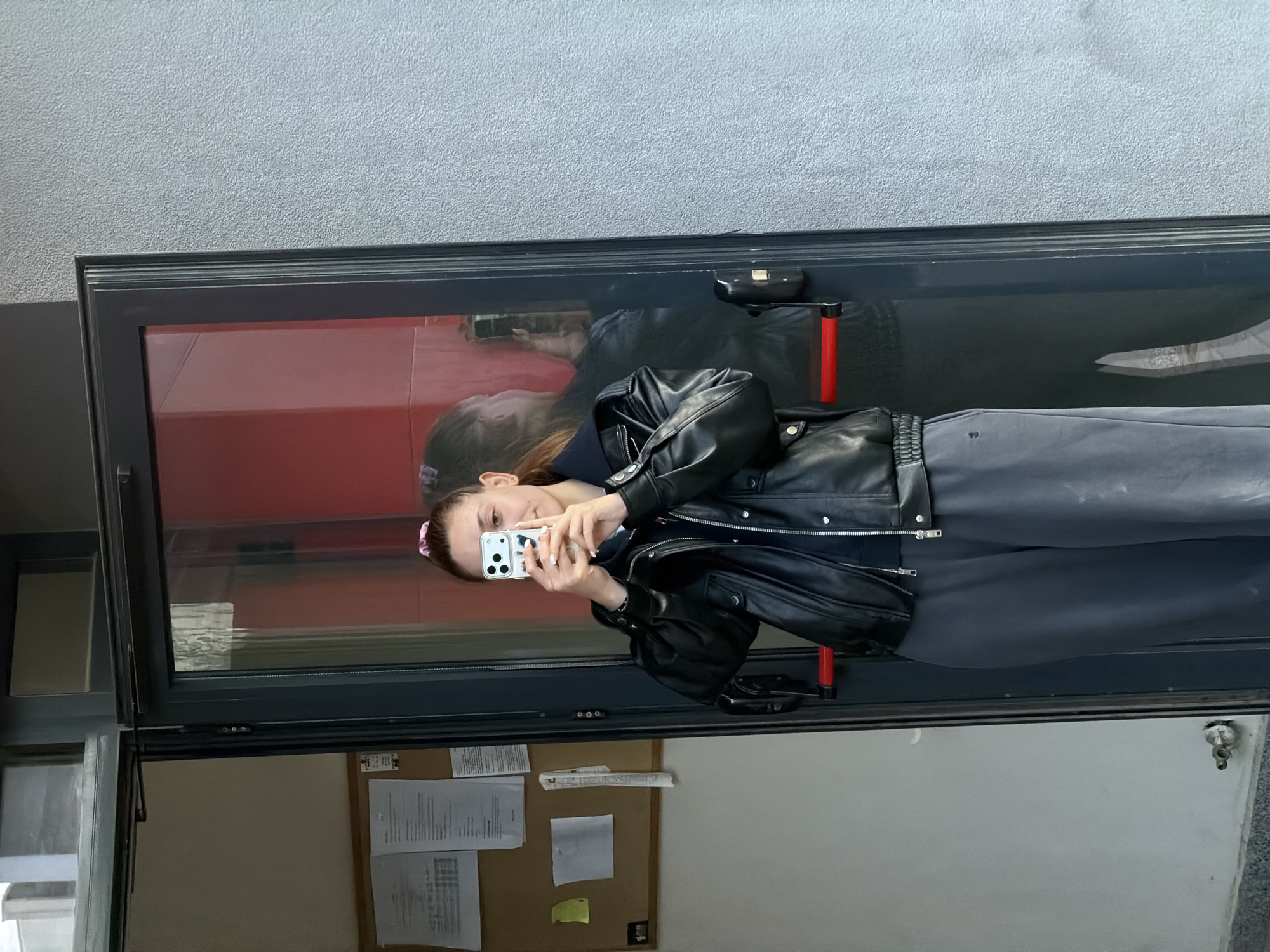 A woman is standing inside a building in Sofia, Bulgaria, taking a photo of herself in a window. The window reflects a red wall inside and gray concrete or a gray painted wall outside. The woman is wearing a black leather jacket and gray pants. She's holding a white smartphone. Behind the window is a bulletin board with papers attached. The wall behind the board is beige.
