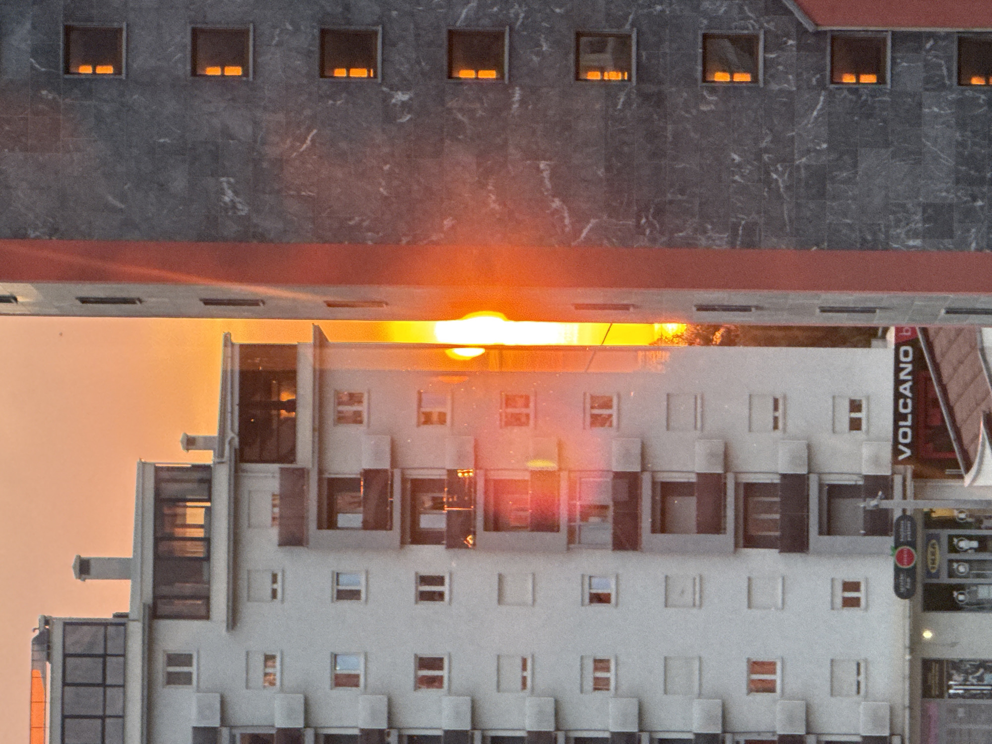 The image depicts a sunset over the city of Bar, Montenegro. Two buildings are visible, seemingly reflecting the scene. The upper building has a grey, textured facade with regularly spaced windows exhibiting orange reflections, suggesting interior lighting or sunset reflection. A red-colored band runs horizontally along the base of the upper building. The lower building, appearing white or light-colored, also features numerous windows and a geometric, textured facade. Some windows reflect the sunset, while others appear dark. A sign reading "VOLCANO" vertically is visible on the right side of the lower building. A traffic light and what appears to be an IKEA sign are attached near it. The sky in the background is predominantly orange, indicating the time of day. The buildings are angled in such a way that they mirror one another across the horizon.