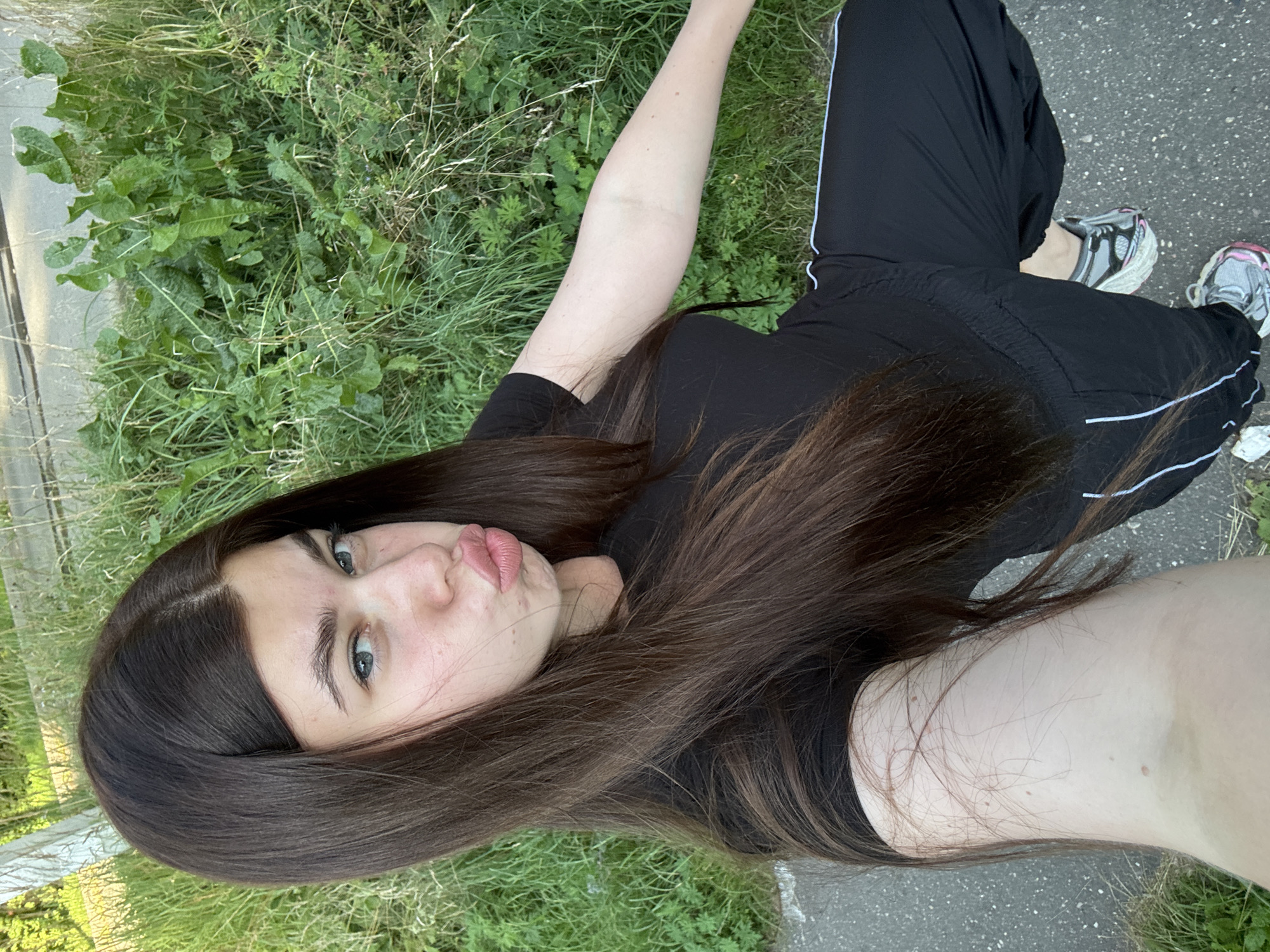 A young woman with long, dark brown hair and light blue eyes is taking a selfie outdoors. She is wearing a black t-shirt and black athletic pants with a white stripe. Her lips are pursed in a playful expression. She is standing on a paved surface next to a patch of tall green grass and weeds. The background suggests an urban or suburban environment with some vegetation. The lighting indicates it is daytime.