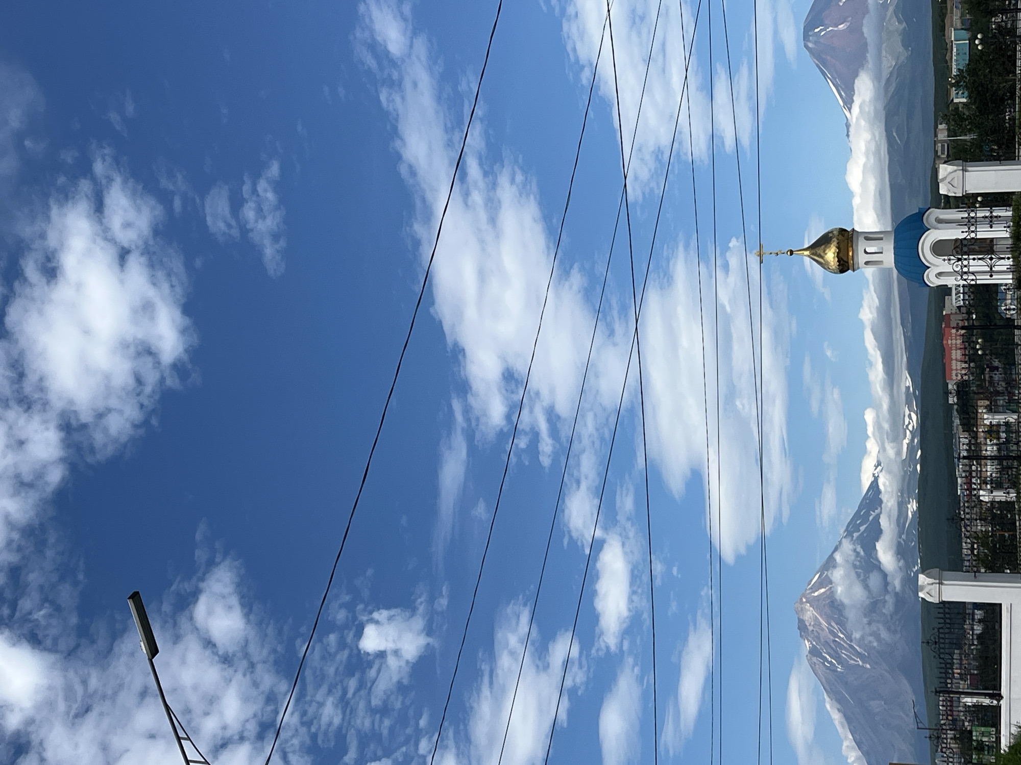 In the city of Petropavlovsk-Kamchatsky, Russia, the scene captures a clear blue sky adorned with scattered white clouds. In the foreground, a street lamp is visible on the left, with several dark power lines stretching horizontally across the frame. A small, white-and-blue Orthodox church with a golden dome and a cross stands in the middle ground. Behind the church, a volcanic mountain with snow-capped peaks rises majestically, partially obscured by clouds. To the right of this prominent volcano, another, smaller volcanic peak is visible, also dusted with snow and ringed by clouds. The lower part of the image shows a low-rise cityscape with buildings and trees, bordered by a black metal fence and white pillars in the immediate foreground. The overall impression is one of a picturesque view in a volcanic region.