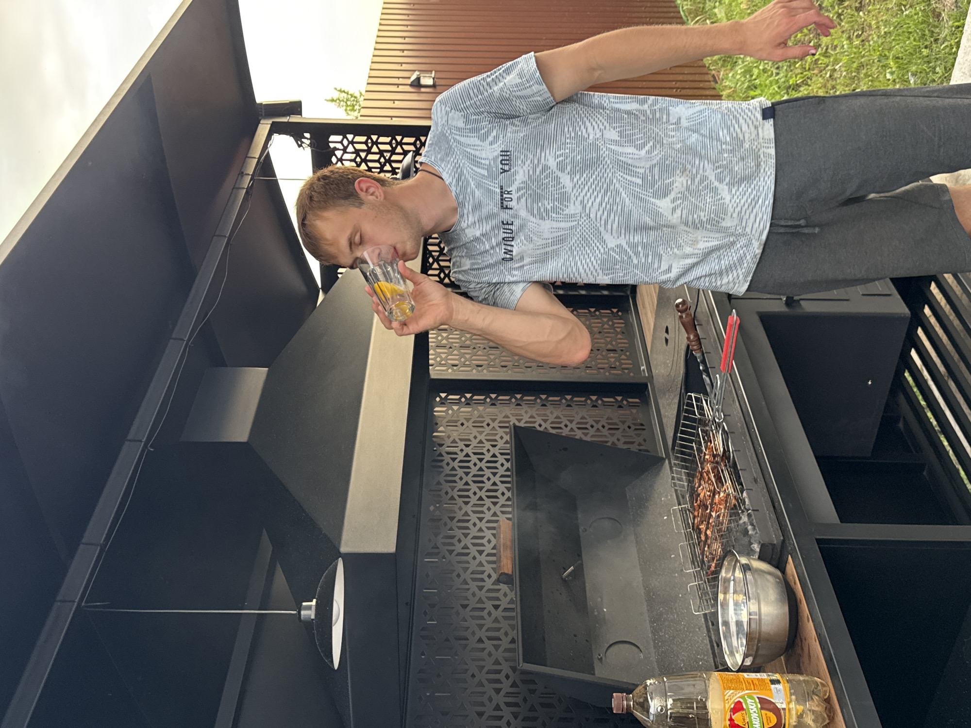 In Starokorsunskaya, Russia, a young man with blonde hair stands beside a black outdoor grill. He is wearing a light blue and white patterned t-shirt and dark gray shorts, and is drinking from a clear glass containing a yellow liquid and ice. The grill is smoking, with meat cooking on a metal rack. To the left of the grill, there is a large plastic bottle of a yellow beverage and a stainless steel bowl. The scene is outdoors, with a wooden fence visible in the background and green grass underfoot. A black metal awning with a hanging light fixture is positioned above the grill.