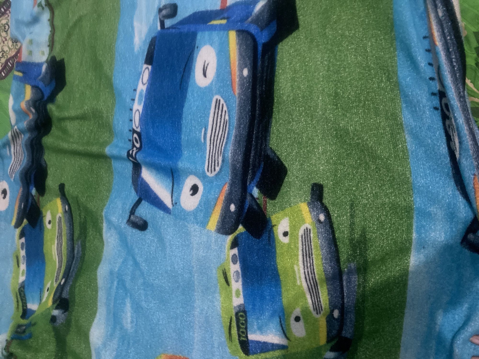 A close-up, overhead shot of a bedsheet with a cartoon design of buses is shown. The bedsheet is primarily blue and green, with the cartoon buses in the foreground. One bus is green with the number "1000" on its front, and the other is blue. The design appears to be from a children's show, possibly "Tayo the Little Bus." The image is taken at an angle, with shadows indicating the lighting source. There is no visible indication of the location in Kalitinggar, Indonesia.