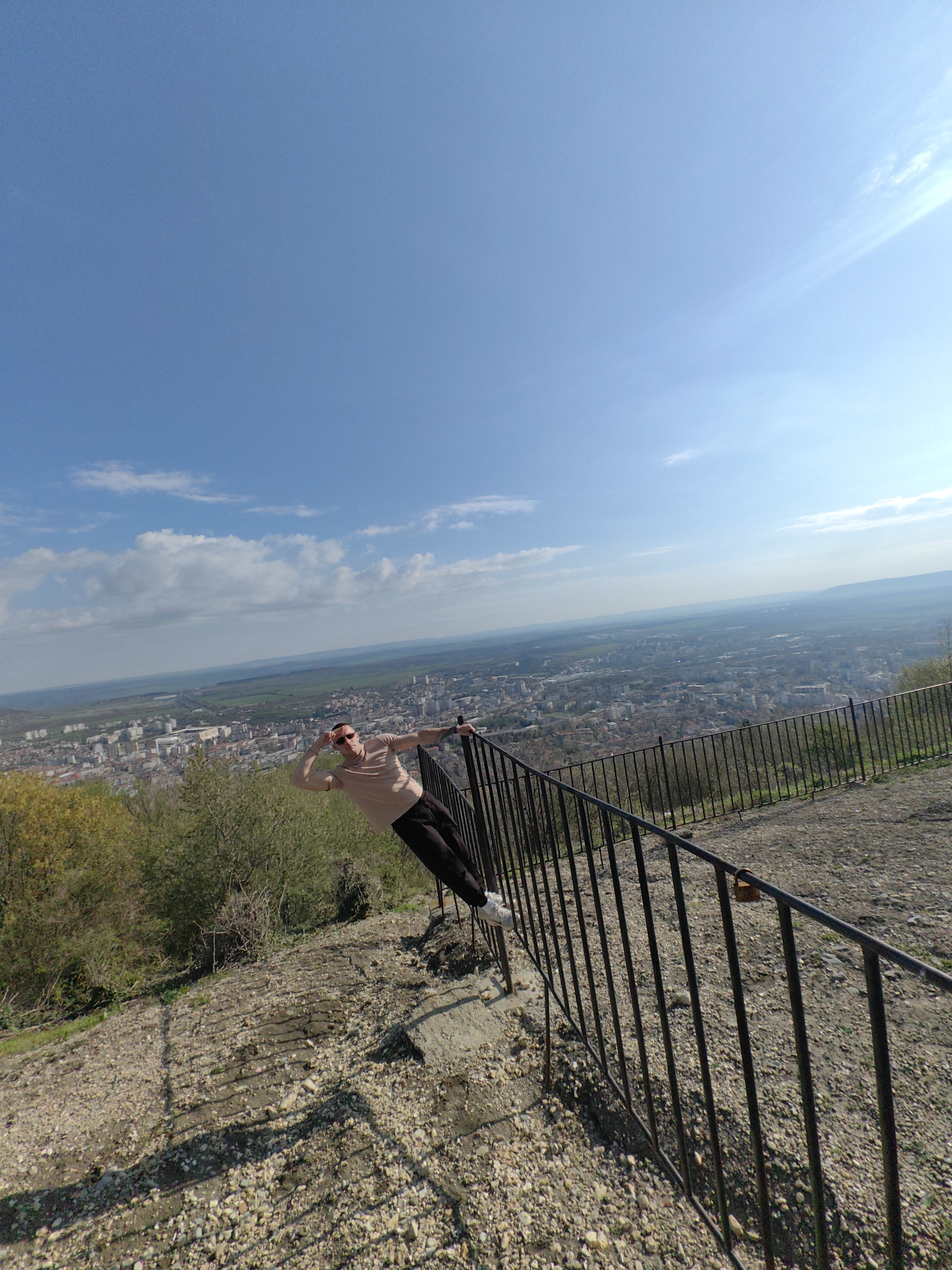 A medium shot of a person leaning on a metal railing on a hillside overlooking Shumen, Bulgaria. The person, a light-skinned male, is wearing sunglasses, a light pink shirt, dark pants, and white shoes. He leans with his right arm on the rail, his left hand up to his forehead. The railing is black, composed of vertical bars. The hillside is covered with small stones and some grass, and in the background there are some trees on the left. Beyond the railing is a view of the city of Shumen with a wide expanse of land and a clear blue sky with some clouds.