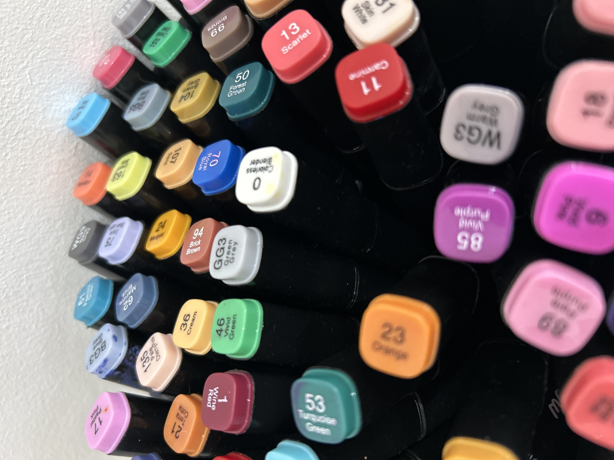 The image shows a close-up view of the tops of many markers, likely arranged in a container or display. The markers have a black body with colored caps that indicate the ink color. The caps are square-shaped with rounded corners and have labels with numbers and color names. The visible labels include "Scarlet 13", "Carmine 11", "Vivid Purple 85", "Turquoise Green 53", "Orange 23", "Wine Red 1" and "Forest Green 50". The markers are grouped together, filling the frame of the image. The background is a plain white surface. The image does not depict any information related to the location in the city of Chaikovsky, Russia.