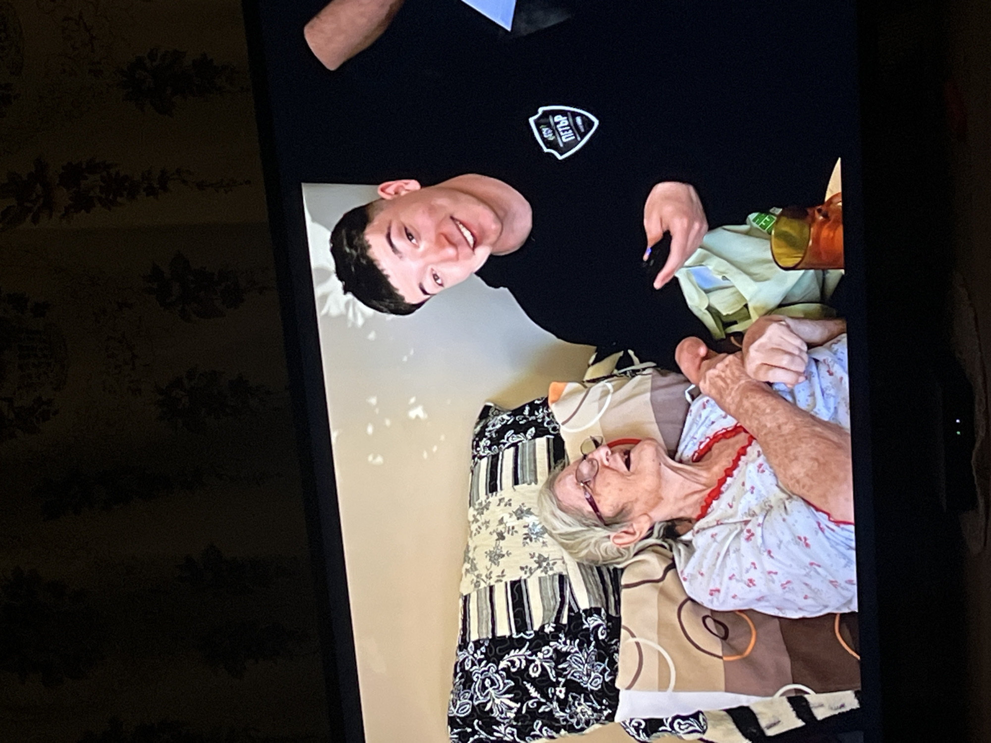 A young man wearing a black t-shirt with a white logo is standing next to an elderly woman lying on a bed. The woman is wearing a light-colored floral shirt with red trim and eyeglasses. The bed has a patterned quilt. A drink in a glass is nearby. The photograph is displayed on a screen, and a wall with patterned wallpaper is visible in the background. The location, Lozarevo, Bulgaria, is known, but there are no specific visual indicators in the image to confirm the location.