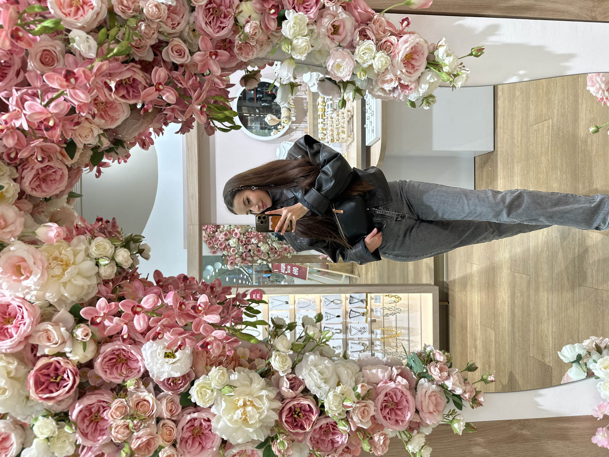 The image displays a young woman taking a selfie in a large, decorative mirror. The mirror's frame is extensively adorned with a dense arrangement of artificial flowers, primarily pink and white roses, peonies, and pink orchids, forming a visually rich border around the reflection.
The woman has long, dark hair and is dressed in a black leather jacket, a dark top, and grey jeans. She is holding a smartphone to capture her image, appearing calm with a gentle smile. Her reflection shows her from the waist up.
The mirror reflects the interior of what appears to be a retail store or boutique. Behind the woman, several display cases are visible, showcasing various jewelry and accessories such as necklaces and earrings on stands, with both gold and silver tones. The store features light-colored walls and a light wooden floor. In one of the reflected display areas, a prominent sign with the Russian text "АКЦИЯ" (meaning "SALE" or "PROMOTION") and the number "690" is visible, along with a partial "360" on another tag. A smaller, round mirror is also reflected in the background. The setting is well-lit, suggesting an indoor retail environment during operating hours.