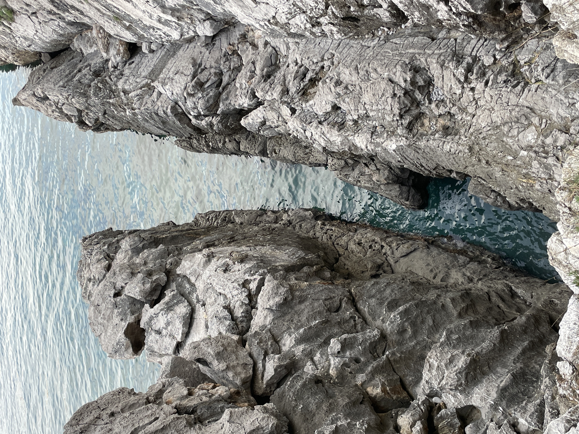 The image displays a deep, narrow natural rock channel situated on the coast of Budva, Montenegro. Two prominent, rugged rock formations, predominantly grey in color with varying textures, flank this channel. The rock surfaces exhibit extensive weathering, characterized by numerous fissures, sharp edges, and undulating, layered patterns indicative of erosion. Seawater, displaying a greenish-blue tint with visible surface ripples, fills the channel and extends into the background. The water within the confined space appears darker due to depth and shadows, contrasting with the lighter, more open water beyond. No individuals, man-made structures, or specific activities are present in the depicted scene, which emphasizes the raw, natural interaction between rock and sea.