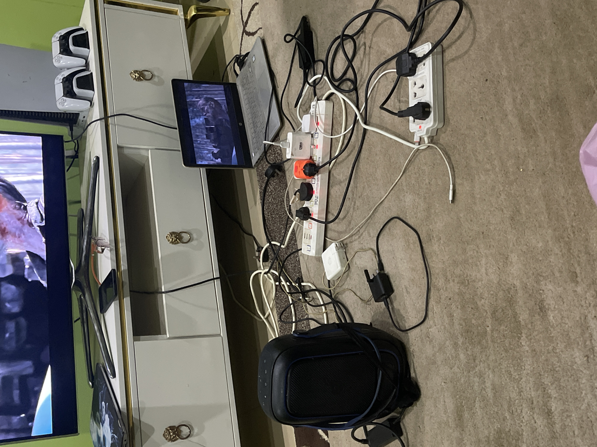 The image shows a cluttered indoor space. A television is on the left, next to a white cabinet with gold-colored knobs. On top of the cabinet are two white game consoles. A laptop sits on top of the cabinet as well. A power strip is plugged into another power strip on the floor. Several electronic devices are plugged into the strips. There is a VR headset on the floor. The location is likely Abuja, Nigeria. The scene suggests a technology-heavy entertainment setup. There are no people visible.