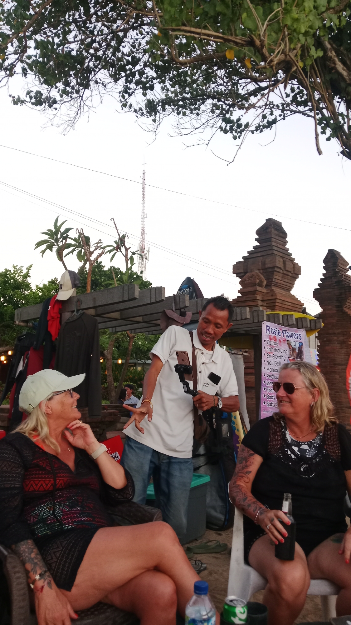 In Kuta, Indonesia, two women and a man are present in an outdoor setting, likely near a beach or street market. The man, wearing a white polo shirt and jeans, is actively engaged, holding a smartphone on a gimbal and gesturing while speaking. One woman, wearing a black patterned dress and a light-colored baseball cap, is seated and looking towards the man, appearing to listen or interact. She has tattoos on her arms and wears glasses. The second woman, also seated and wearing a black top, holds a dark bottle and smiles, also looking in the direction of the man. Behind them, a stand displays services such as "Rosie Flowers," "Tattoo," "Hair Braiding," and "Nail Gel." The background shows foliage, a large transmission tower, and decorative stone structures, suggesting a tropical environment. Clothing is hung on a rack, and various bottles and cans are visible on a table in the foreground.