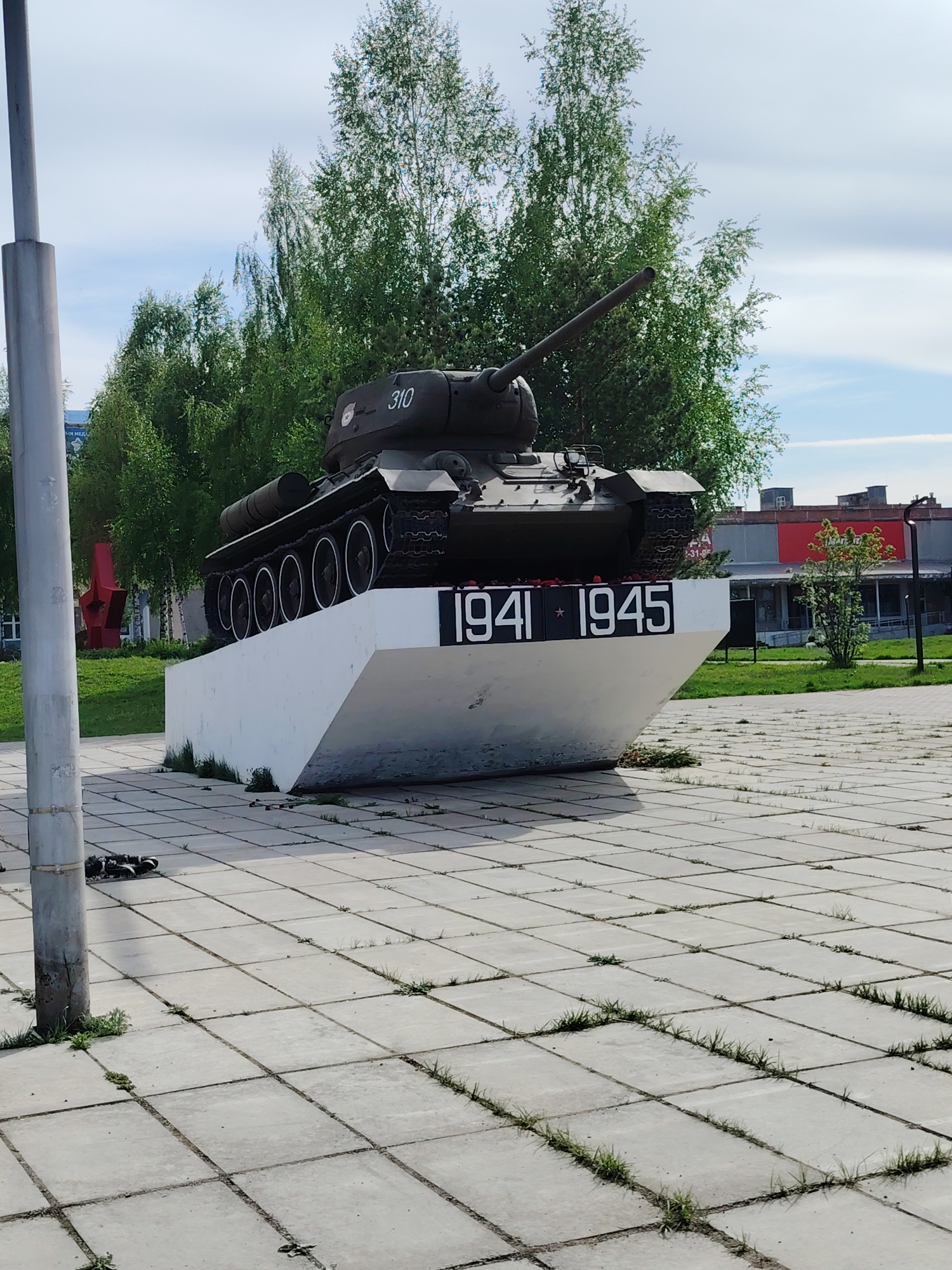 Here is a description of the image:
The image shows a T-34 tank memorial in the city of Марковский, Russia. The tank, painted in dark green, is mounted on a white concrete pedestal bearing the years "1941" and "1945", marking the period of the Great Patriotic War (World War II). The tank is positioned facing the right of the frame, with its gun barrel extending towards the upper right.
The memorial is located in a paved plaza with square concrete tiles. Sparse grass grows between the tiles. A metal pole is visible on the left side of the frame.
In the background, there are green trees and a building with a red section. The sky is partially cloudy.